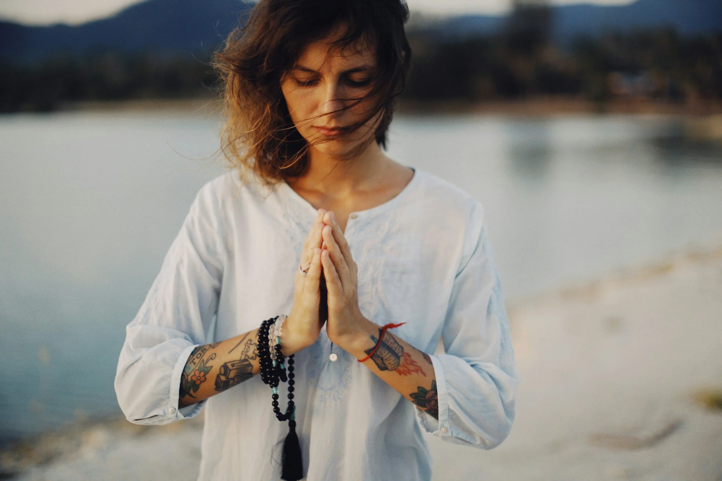 A young woman with dark, wavy hair and tattoos on her arms is standing outdoors near a body of water, holding her hands together in a prayer pose with her eyes closed, appearing to be praying or meditating.