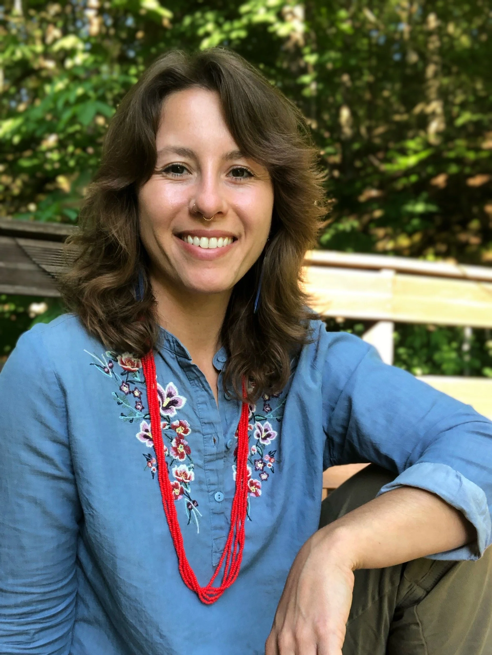 A woman with brown, wavy hair smiling at the camera, wearing a blue embroidered shirt and a red beaded necklace, sitting outdoors with green foliage in the background.