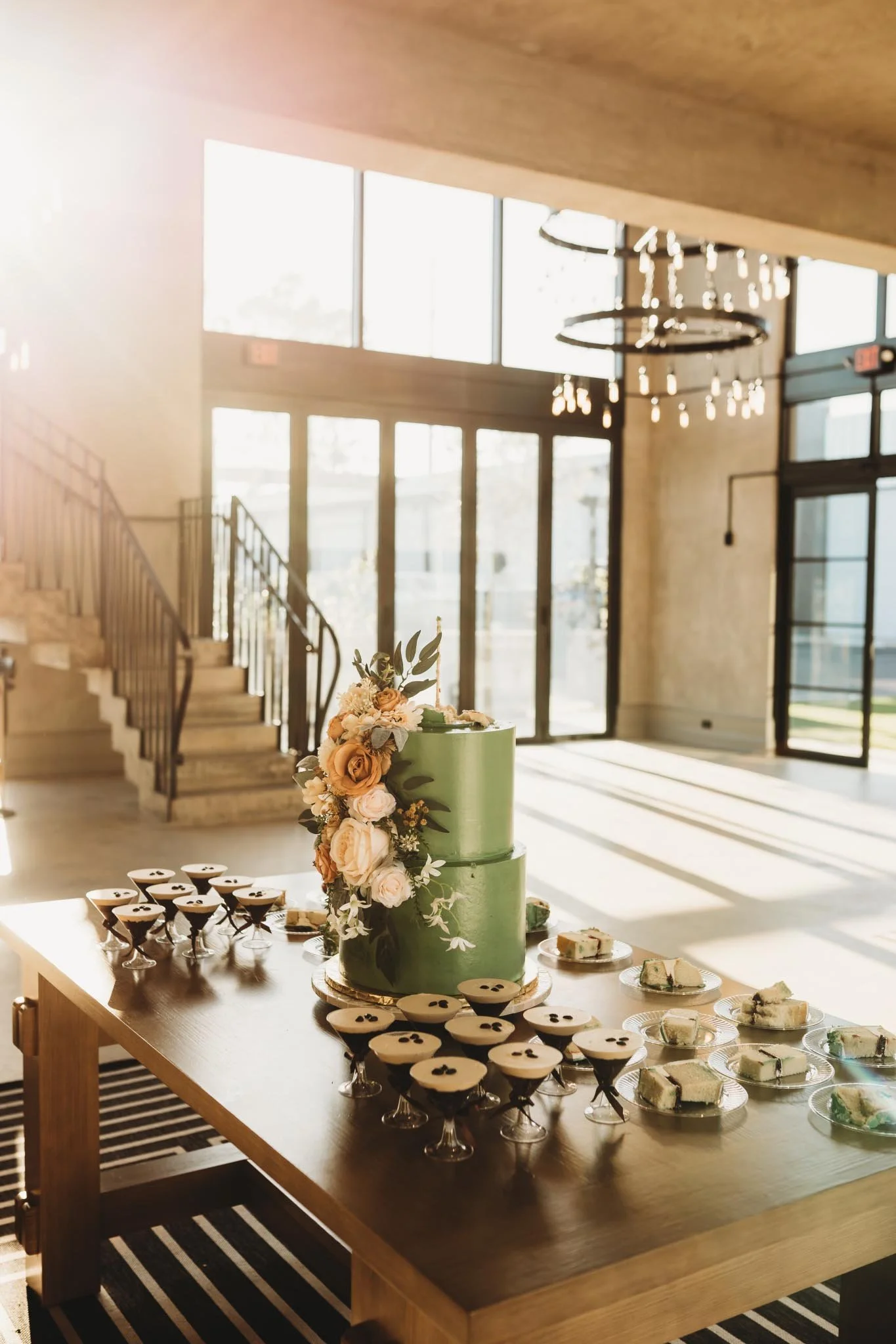 A modern wedding cake on a table with deserts and espresso martinis inside of The Heritage House with a striking golden hour sunlight coming through the large windows
