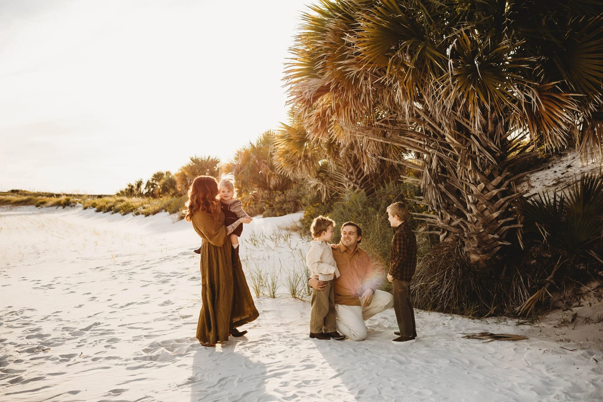 A family of five enjoying a day at the beach, with three children and two adults, standing near a large palm tree on sandy dunes during sunset.