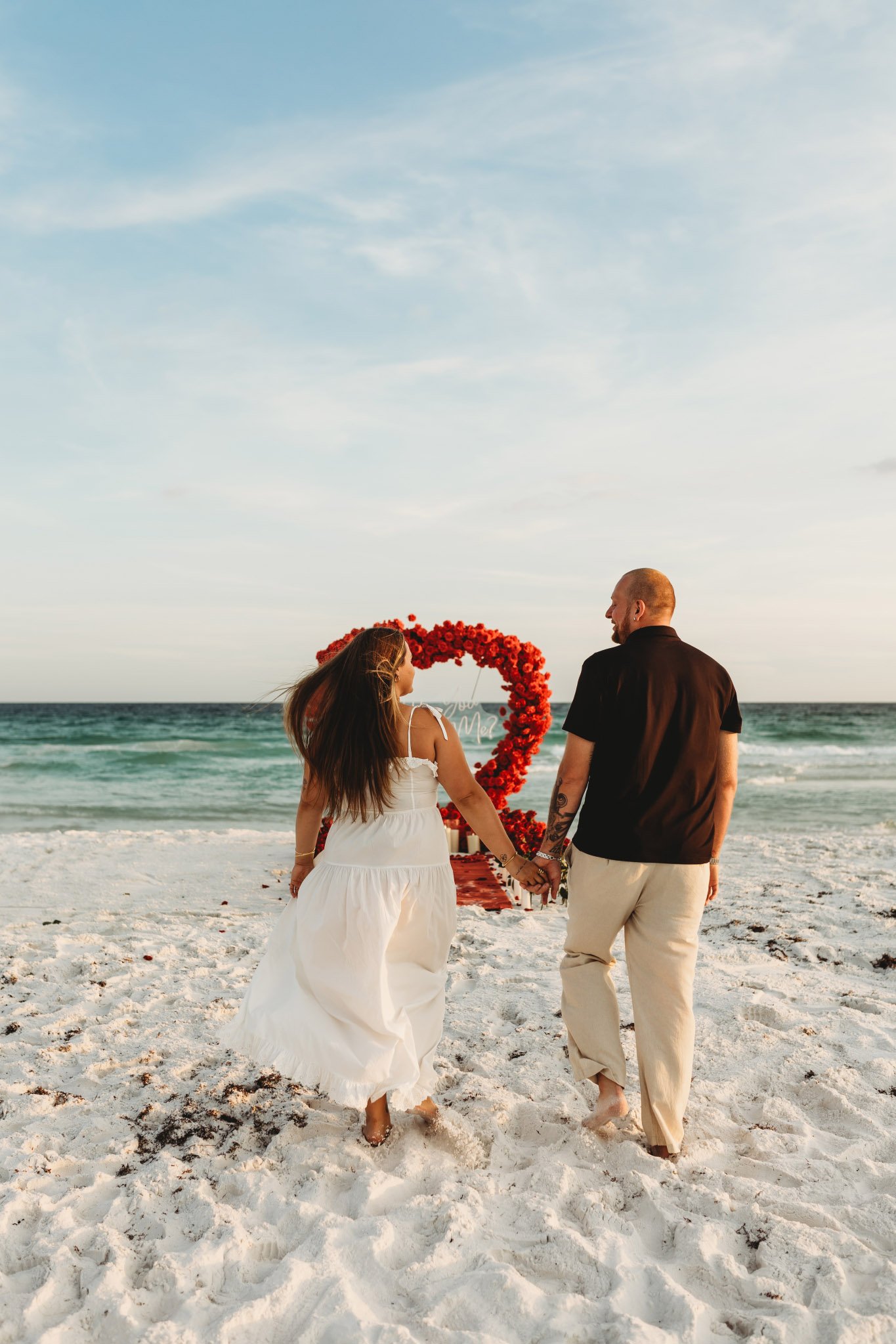 Man and woman holding hands and walking towards a romantic proposal set up on the beach with red roses in the sand