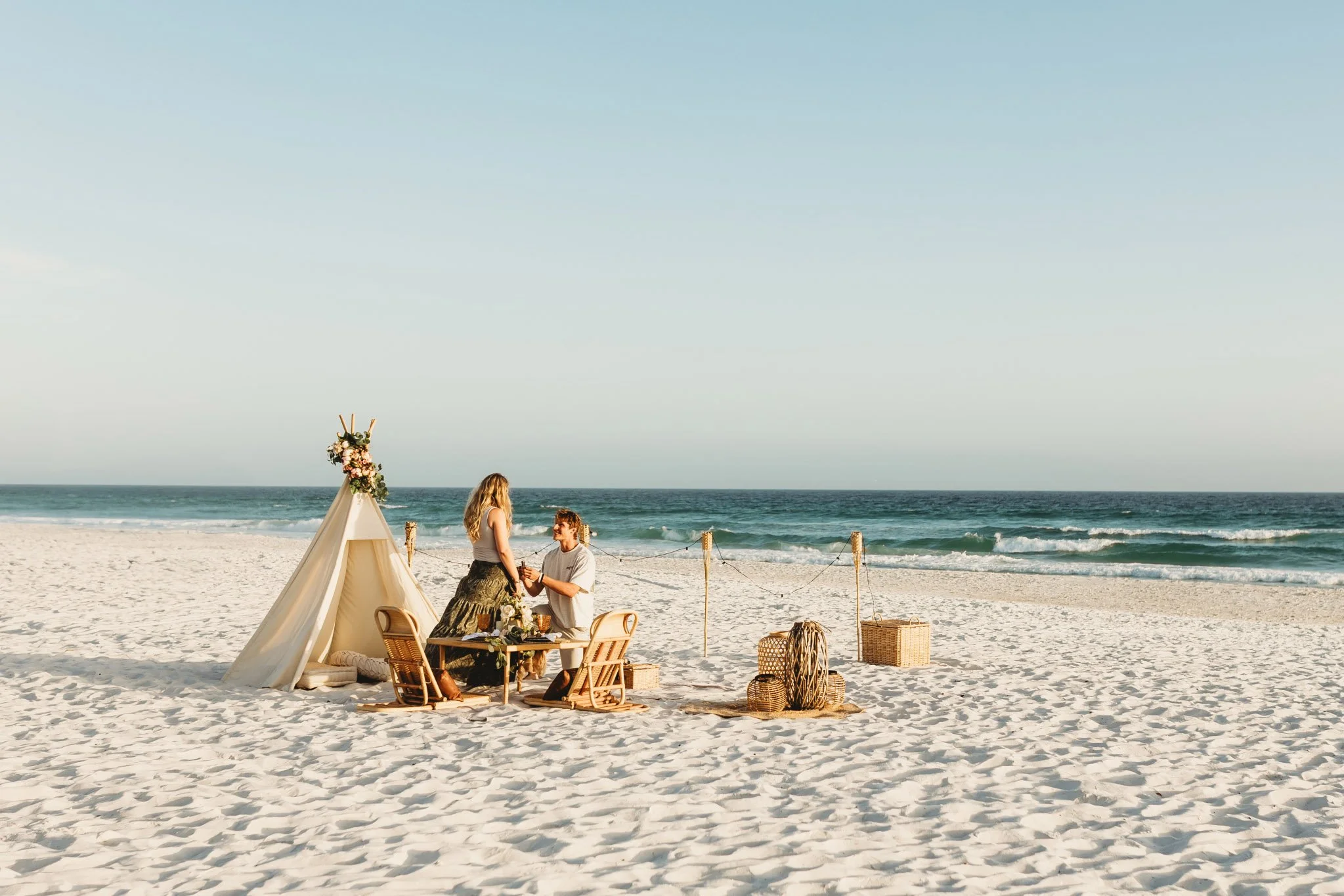 Boho style surprise beach proposal with a tent, chairs, a small table and tiki lights for a private dinner. A man is on his knee in front of a girl.