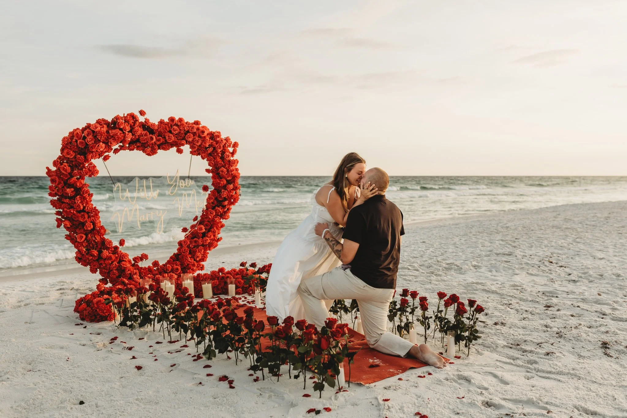 A man on one knee kissing a woman in a white dress on the beach. They are on a red runner in the sand surrounded by dozens of roses and candles. A large floral heart is at the end of the runner with a sign that says "Will you marry me?"