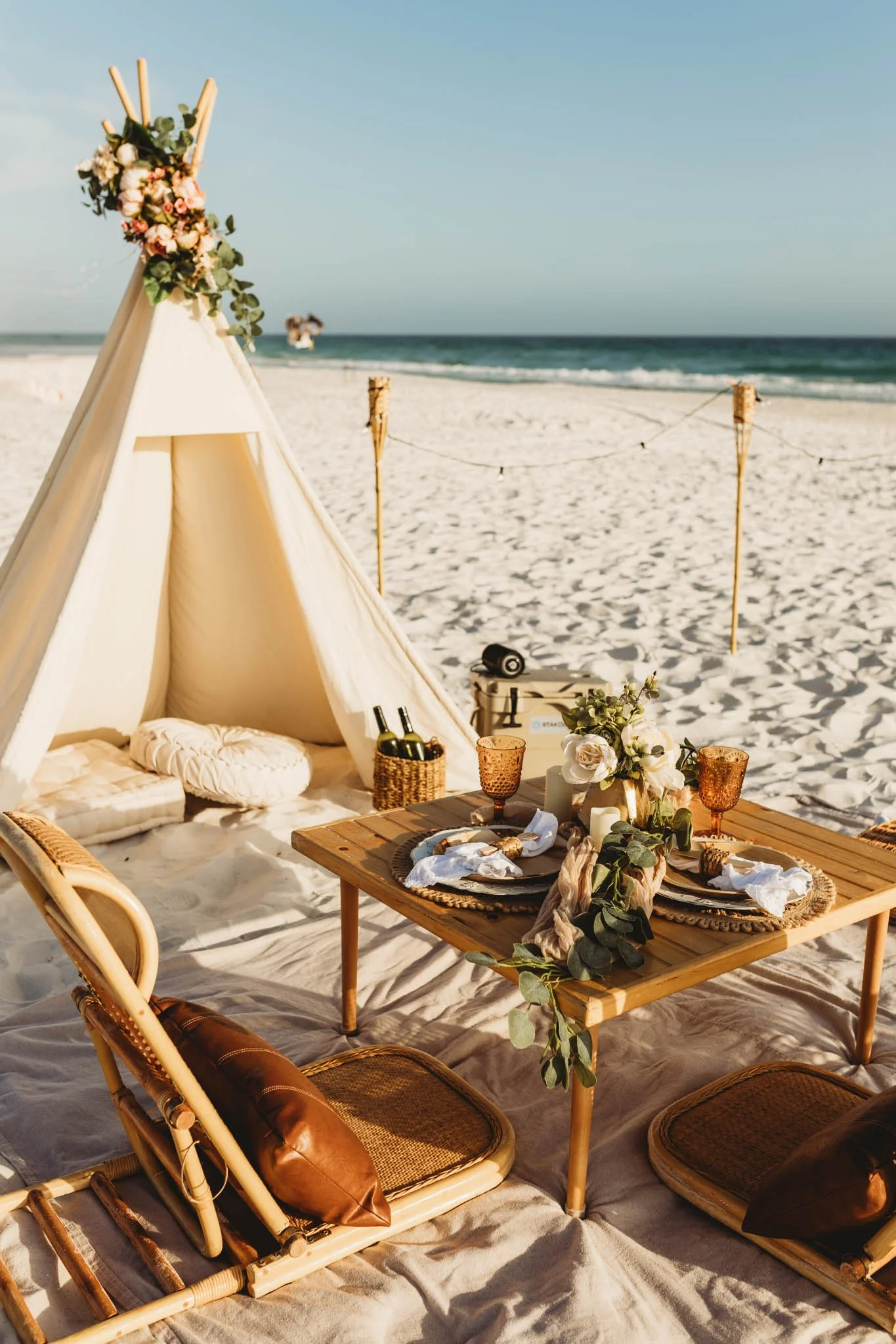 Close-up of a romantic boho style beach picnic with a table, chairs, romantic decor and champagne bottles