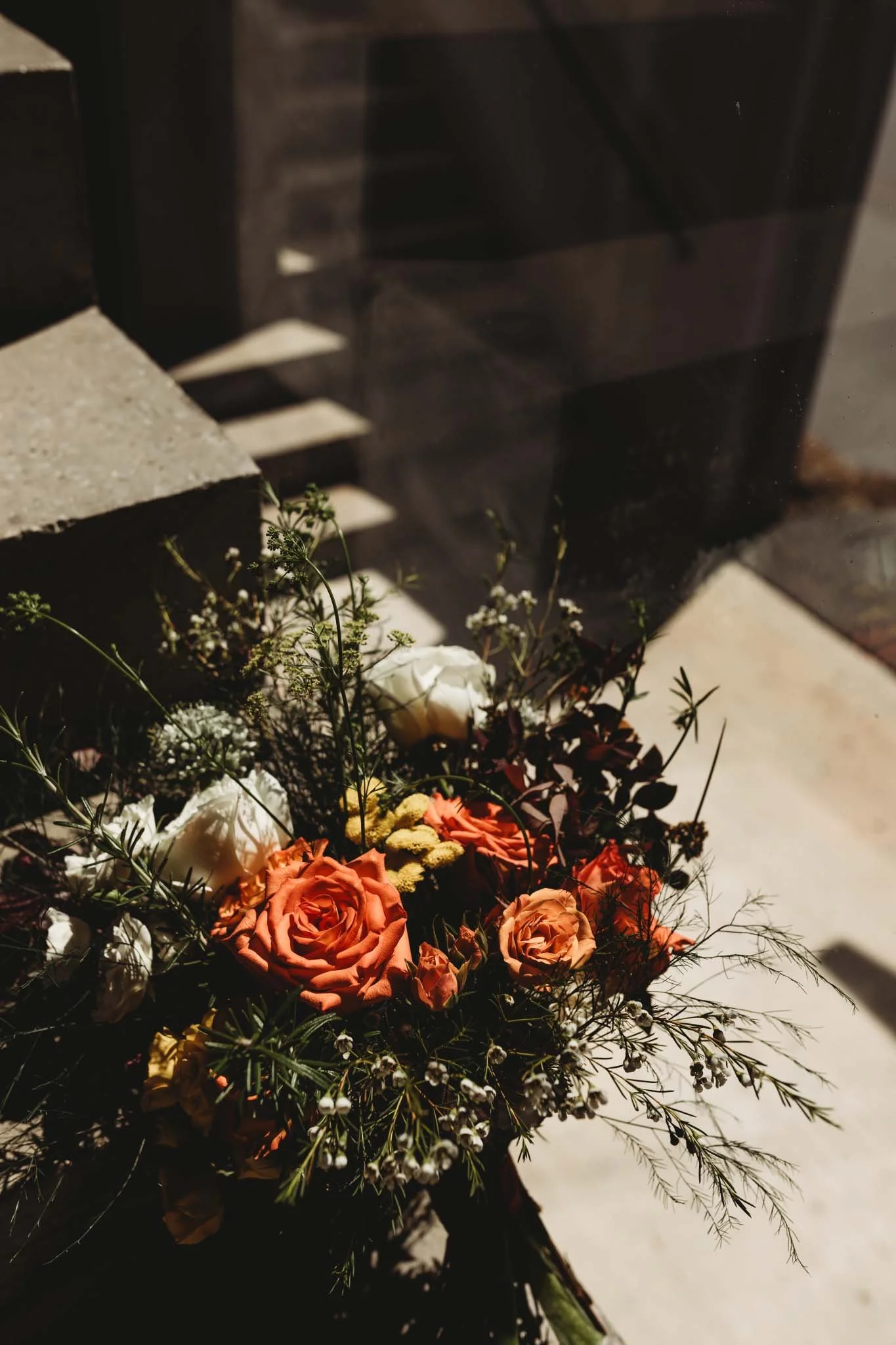 An artistic photo of a bridal bouquet on concrete stairs next to a large window with sharp shadowing