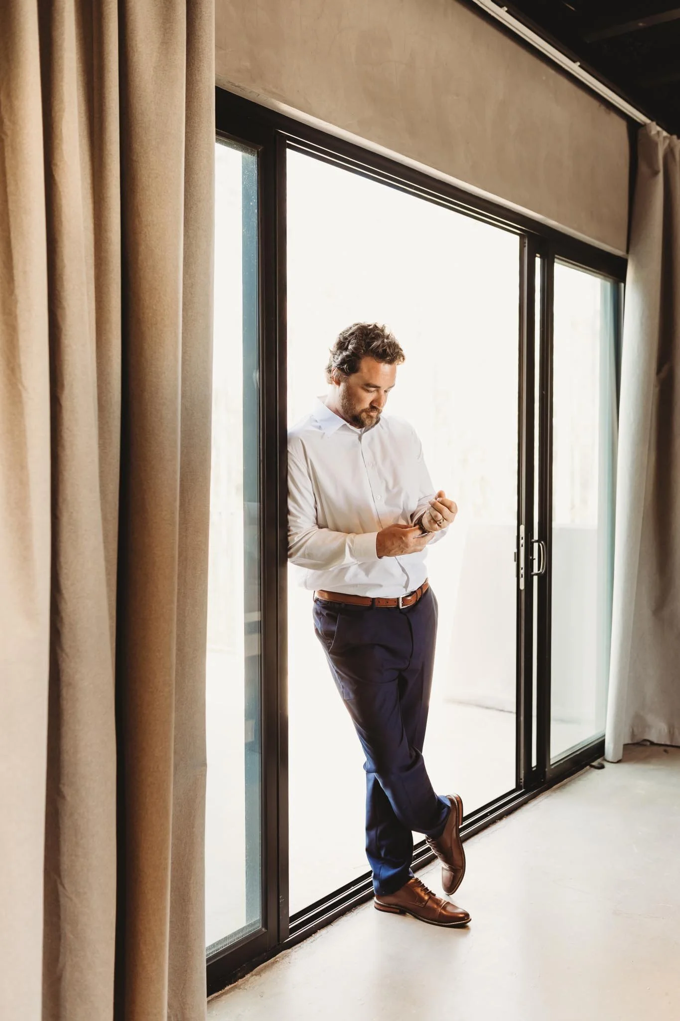 Groom adjusting cufflinks while leaning against a large window in the groom suite at The Heritage House