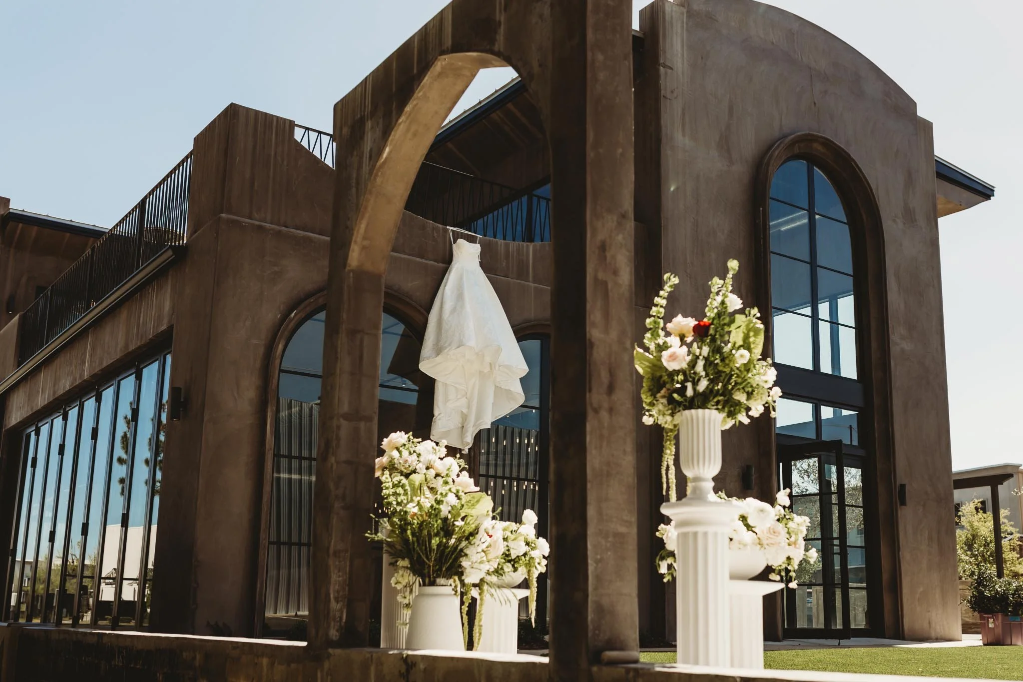A stunning image of a wedding dress hanging on the side of The Heritage House with large floral arrangements below
