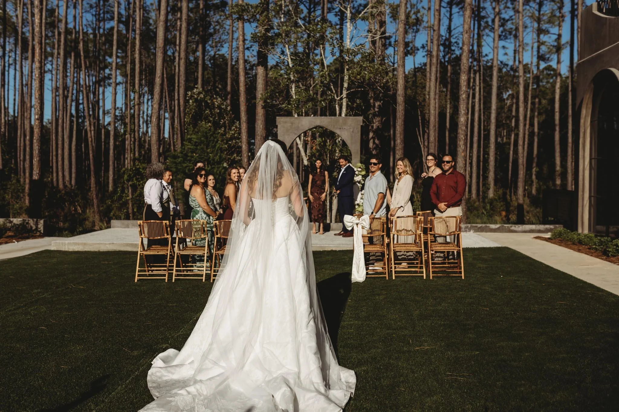 Bride walking down the aisle on the outside lawn. A concrete arch stands at the end in front of tall trees.