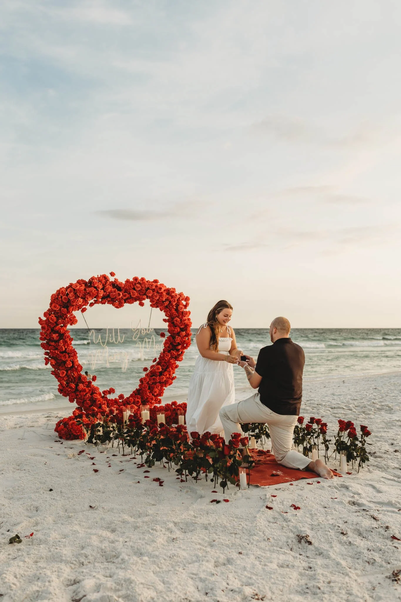 Romantic beach proposal with a man on his knee in front of a girl. They are surrounded by red roses and a large floral heart with a sign that says "Will You Marry Me?"