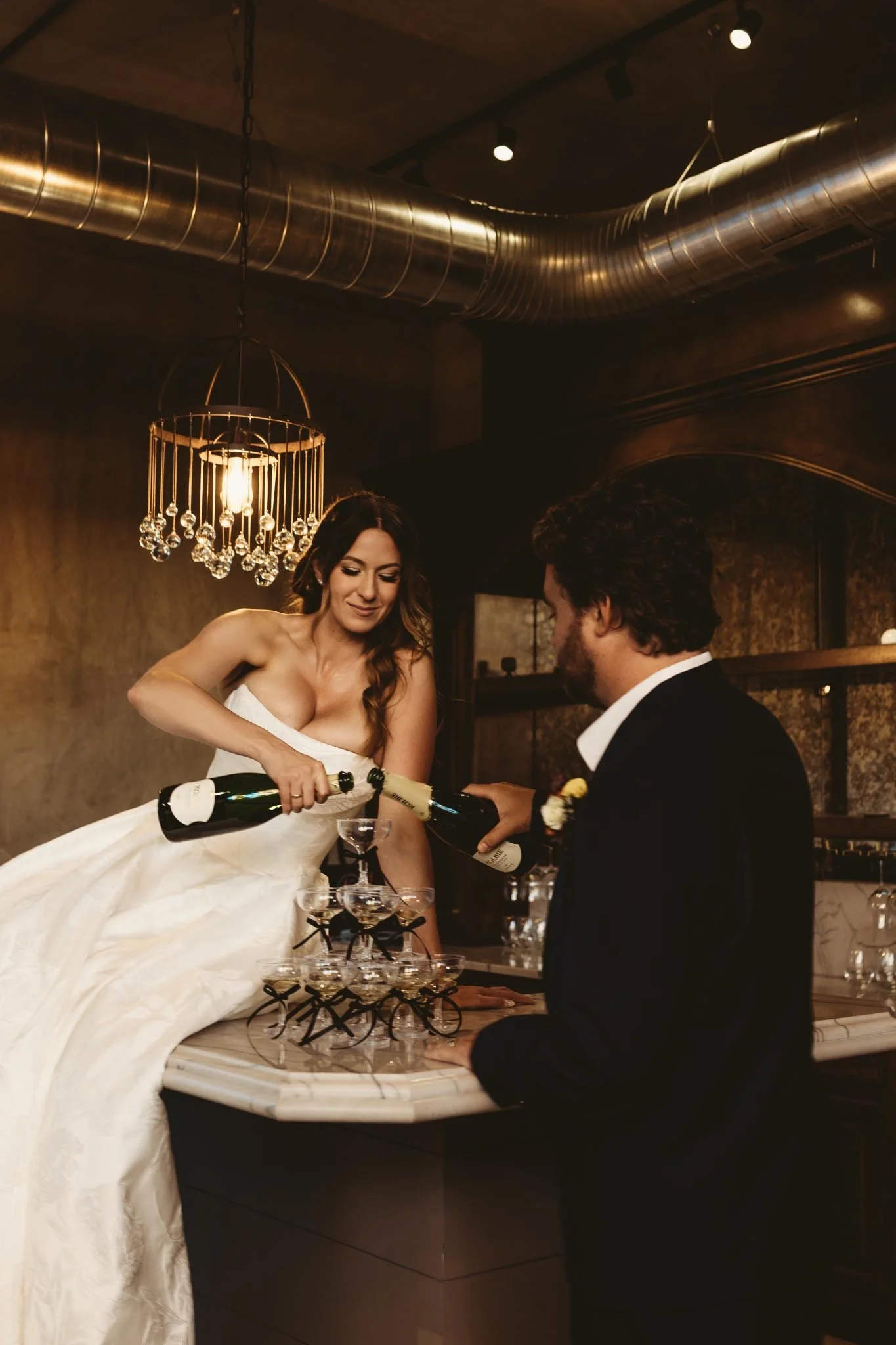 Bride sitting on a bar pouring champagne in a champagne tower with her husband next to her