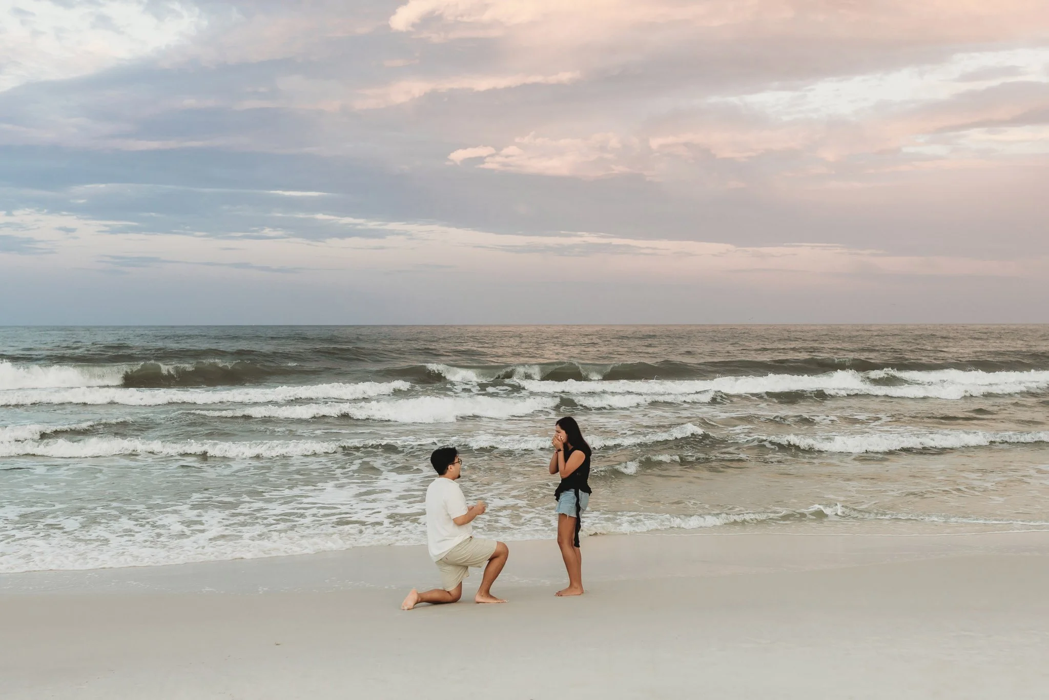 Surprise beach proposal at sunset with a man on his knee and a girl with her hands over her mouth