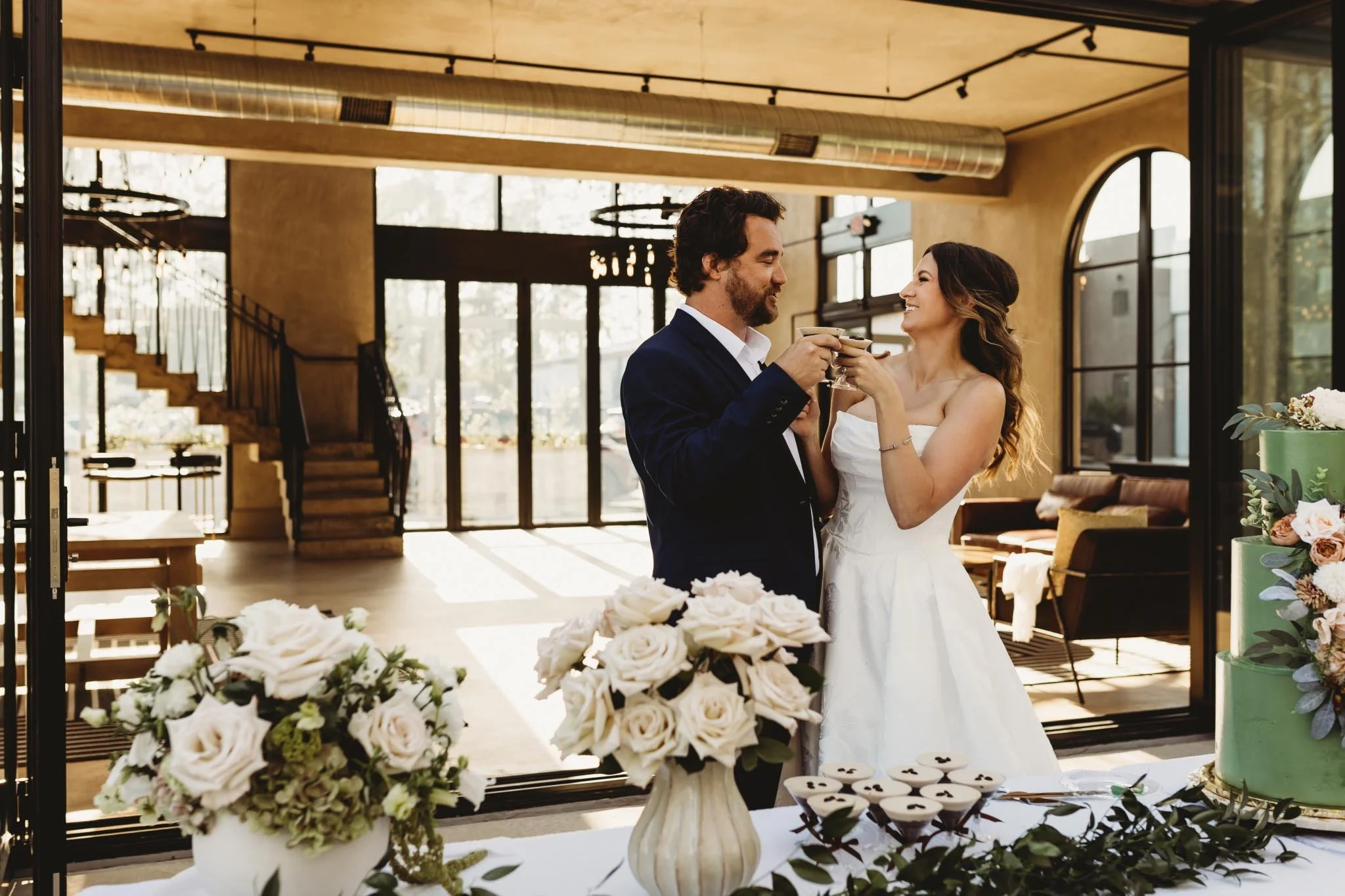 Bride and groom clinking martini glasses in front of The Heritage House. A cake and flower arrangements are on a table next to them.