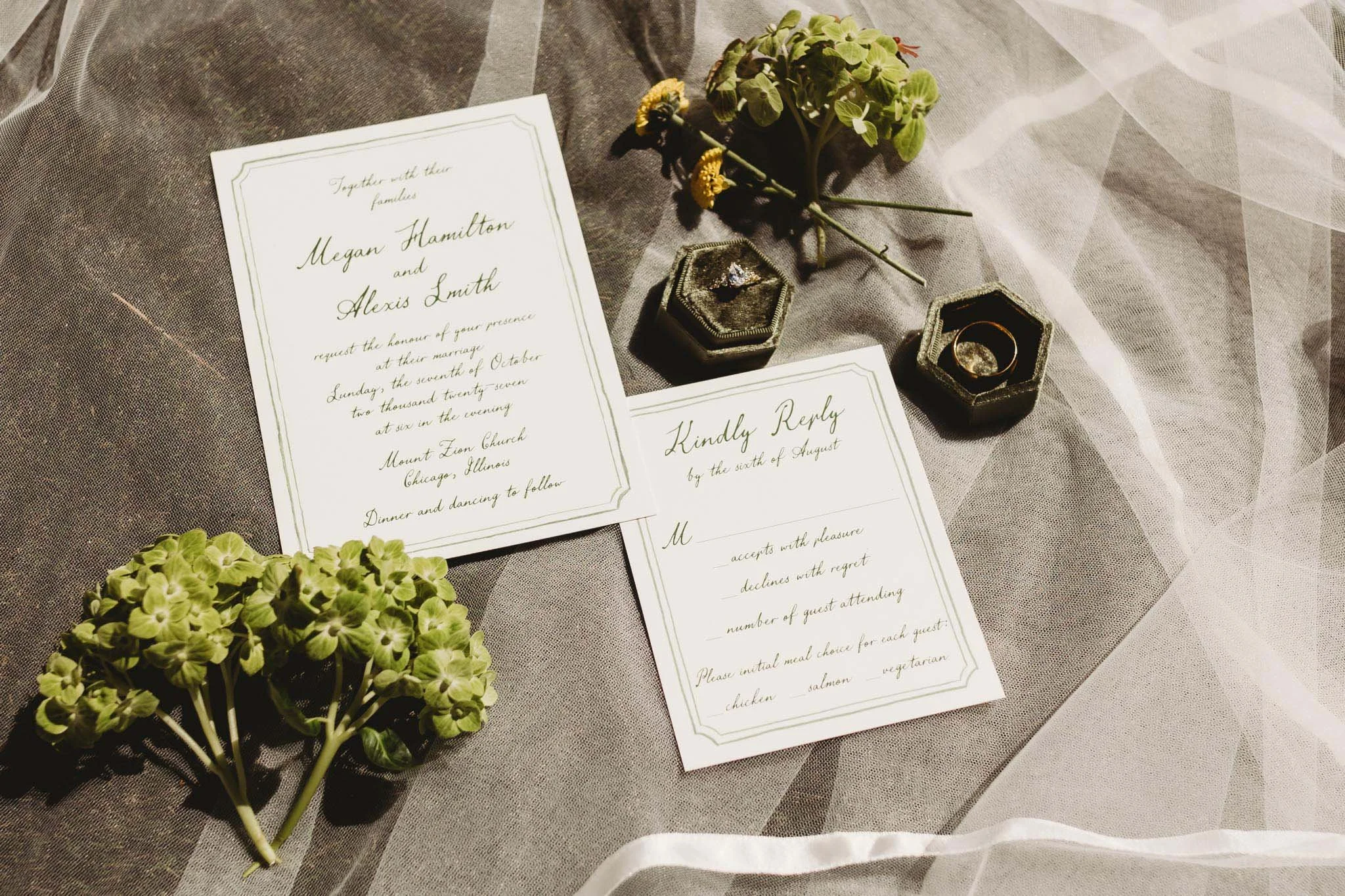Flatlay of wedding invitations, flowers and rings on top of a veil.