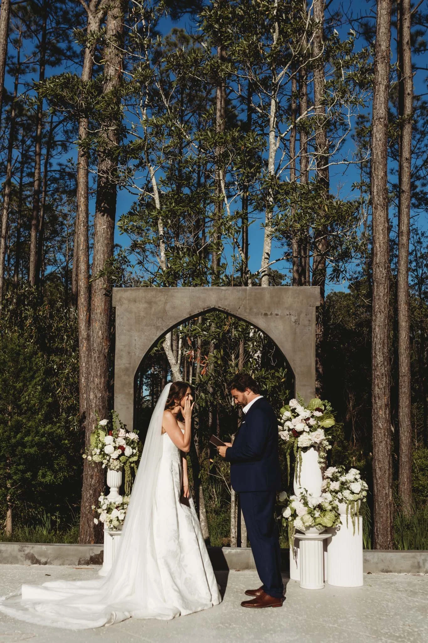bride and groom exchanging vows in front of a modern concrete arch. white flower arrangements surround the set up with tall trees a a backdrop.
