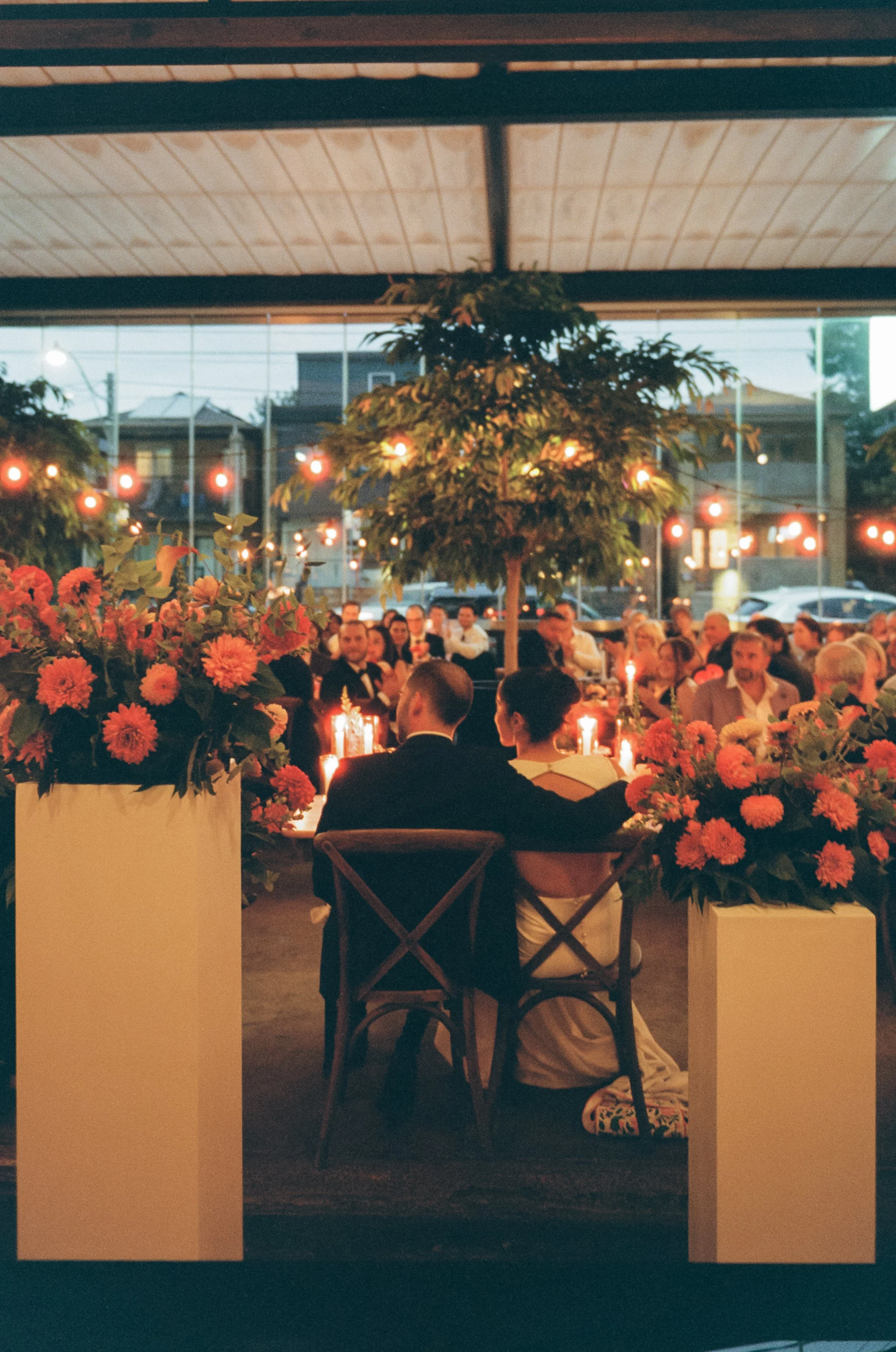 A couple is sitting at a table with candles, surrounded by pink flowers, in a decorated indoor venue with hanging string lights and large windows showing a street view.