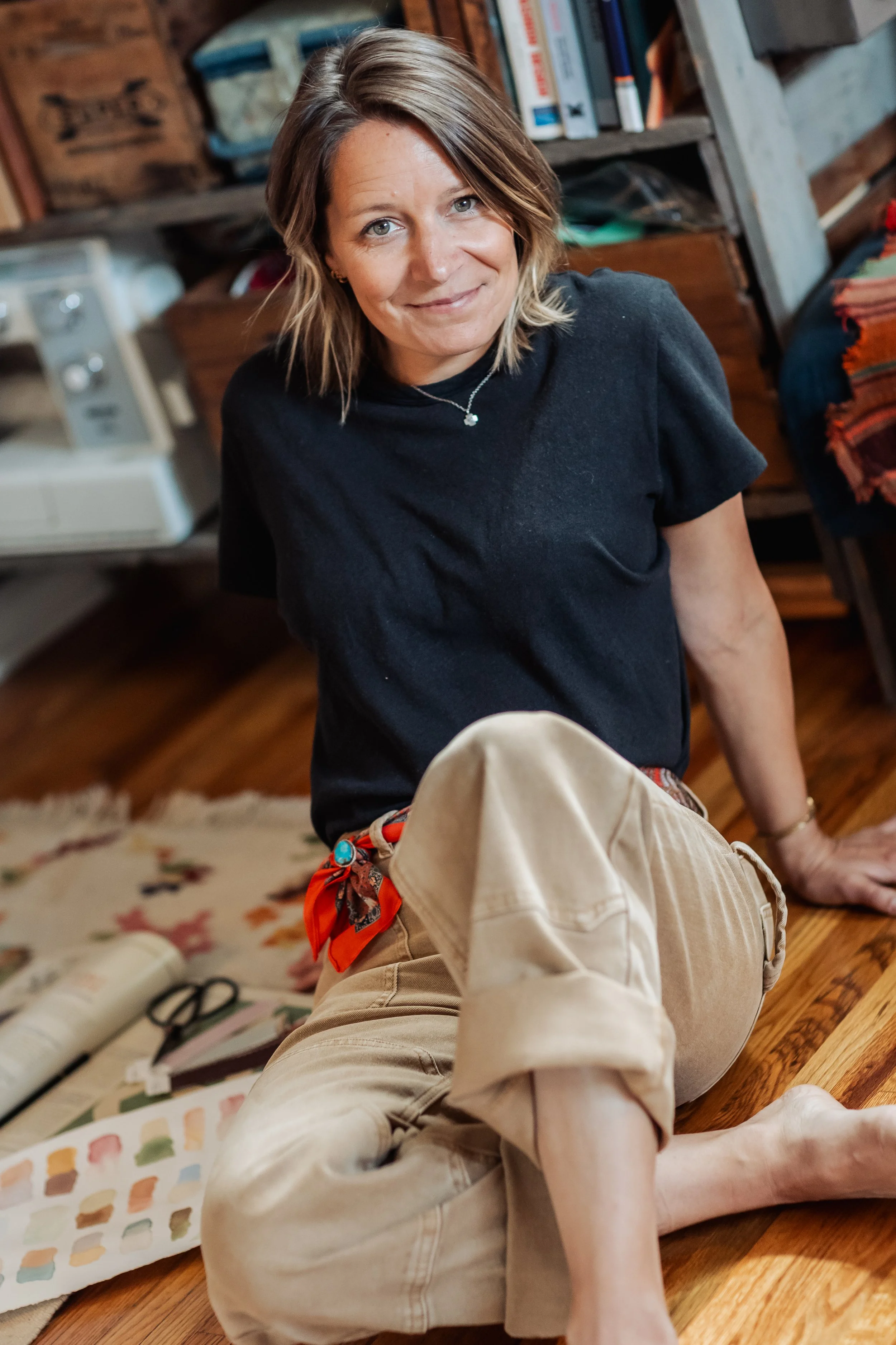 A woman with shoulder-length hair sitting on the floor in a cozy room with wooden shelves and books behind her. She is smiling and wearing a dark t-shirt and a necklace.
