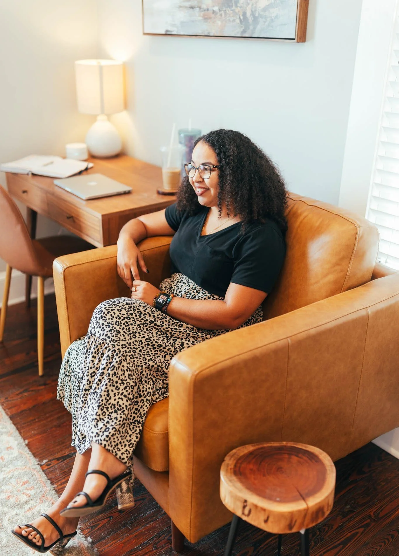A woman with curly hair and glasses sitting on a tan leather couch in a cozy living room, smiling, wearing a black top and leopard print pants, with a laptop and drinks on the wooden desk behind her.