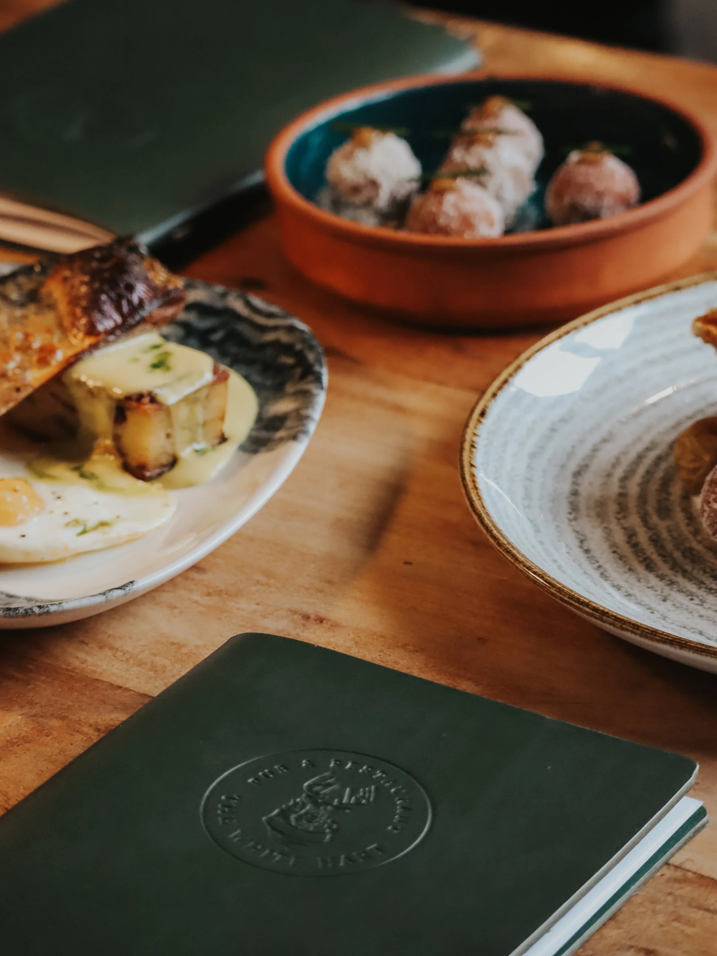 A green menu, several dishes including a bowl of meatballs, a plate with slices of bread, and a dish with eggs and roasted potatoes, on a wooden table.