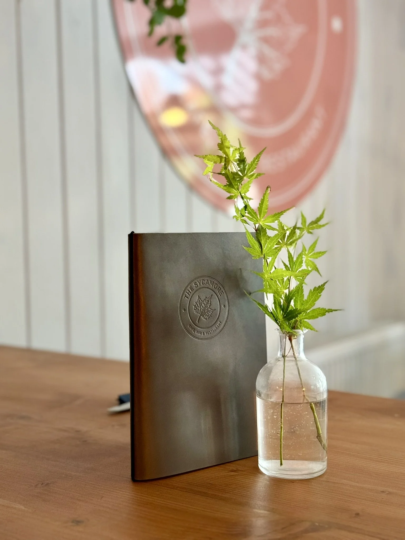 A clear glass vase with water holding a green leafy plant on a wooden table, with a gray menu and a pink decorative plate in the background.