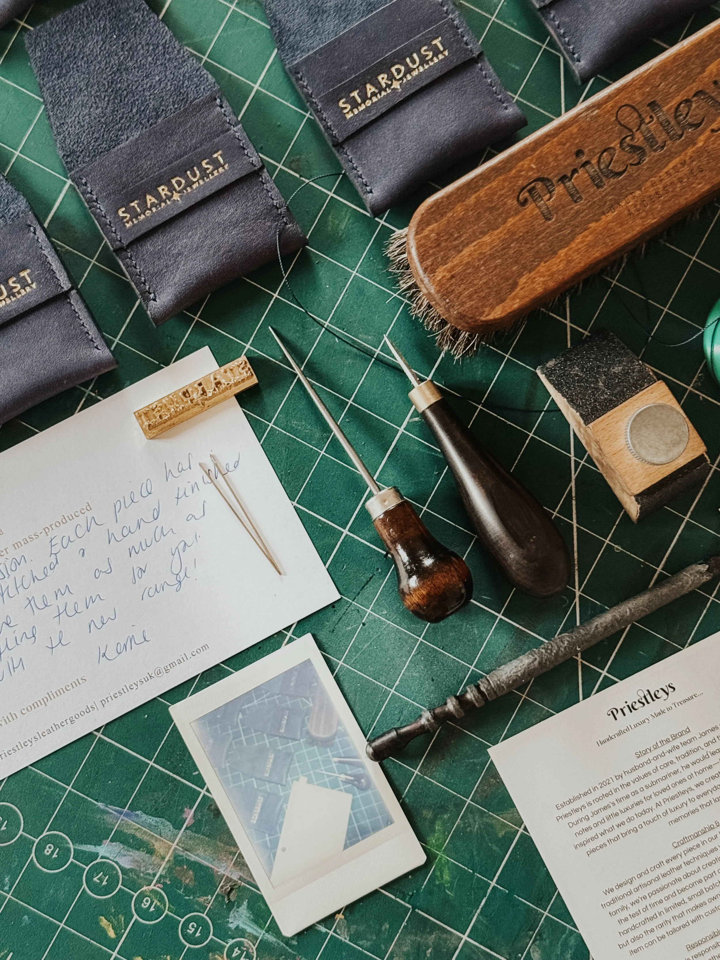 A crafting workspace with leather swatches labeled 'STARDUST' and 'STARMUSE,' various tools including metal needles, a leather punch, a wooden handle detailing a tool with 'Trice,' a piece of paper with handwritten notes, and a photo of a city skyline. The workspace is on a green cutting mat with a grid and measurement markings.