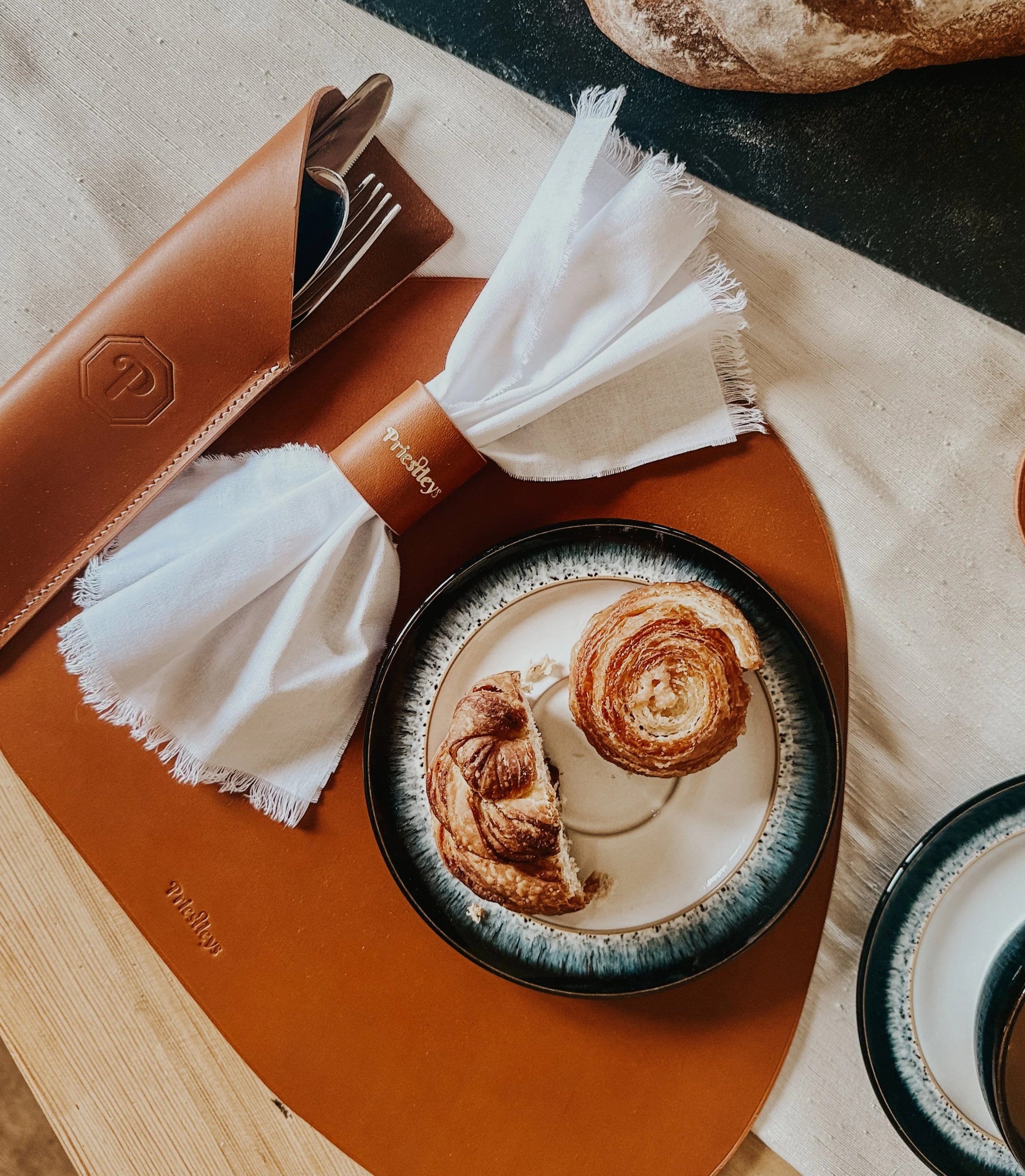 Plate with croissants and pastries placed on a table, with napkins and cutlery in a leather holder nearby.