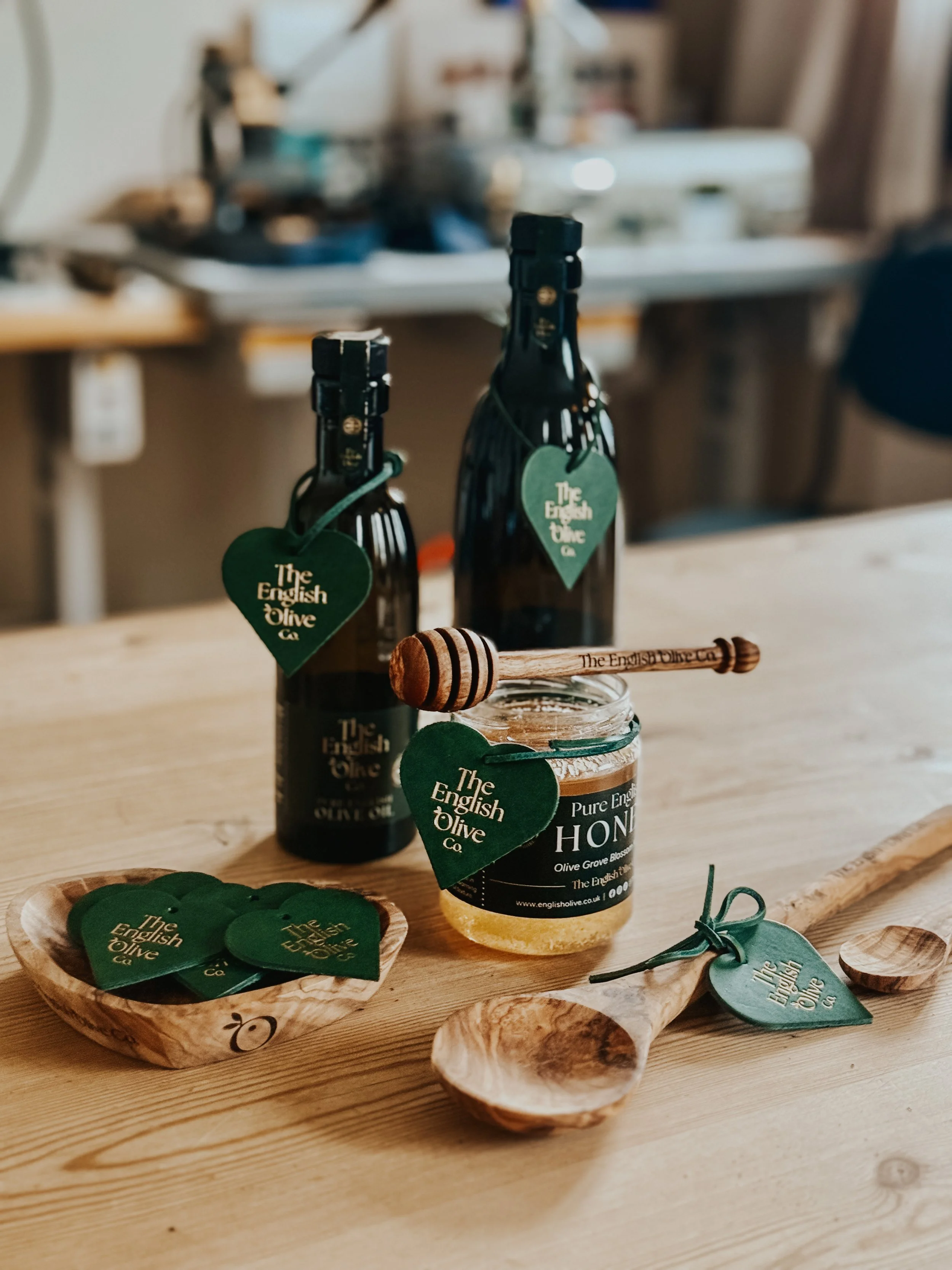 Two bottles of English olive oil with heart-shaped tags, a jar of honey with a wooden honey dipper, and several green heart-shaped tags on a wooden table.