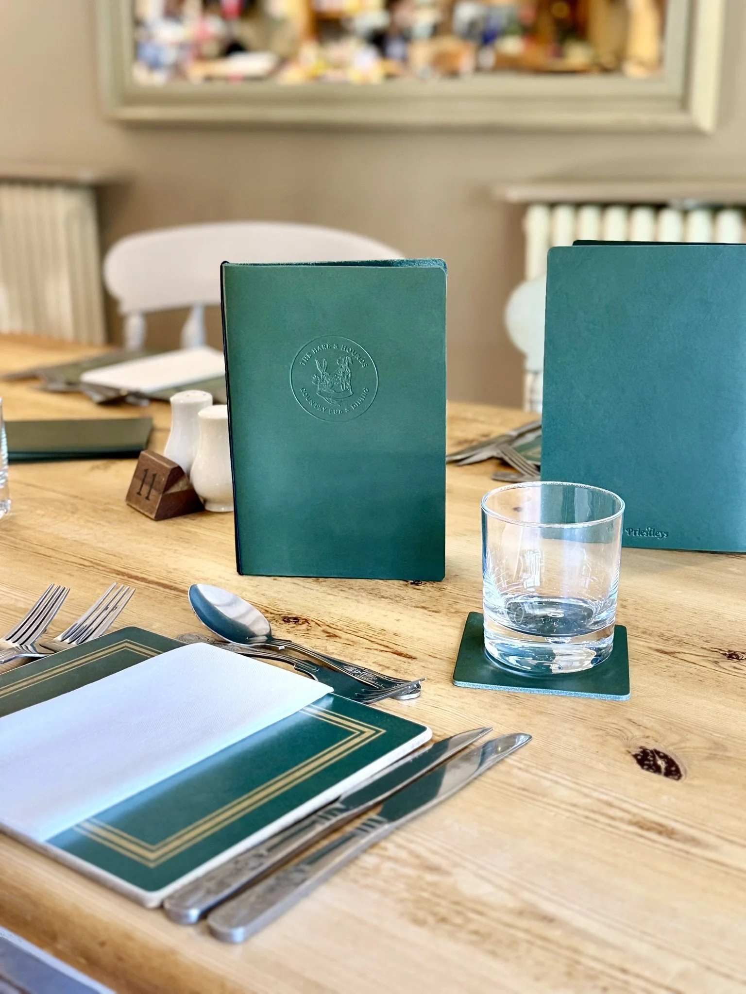 A dining table set with green menus, a glass of water on a coaster, utensils, a white napkin, salt and pepper shakers, and white chairs in the background.