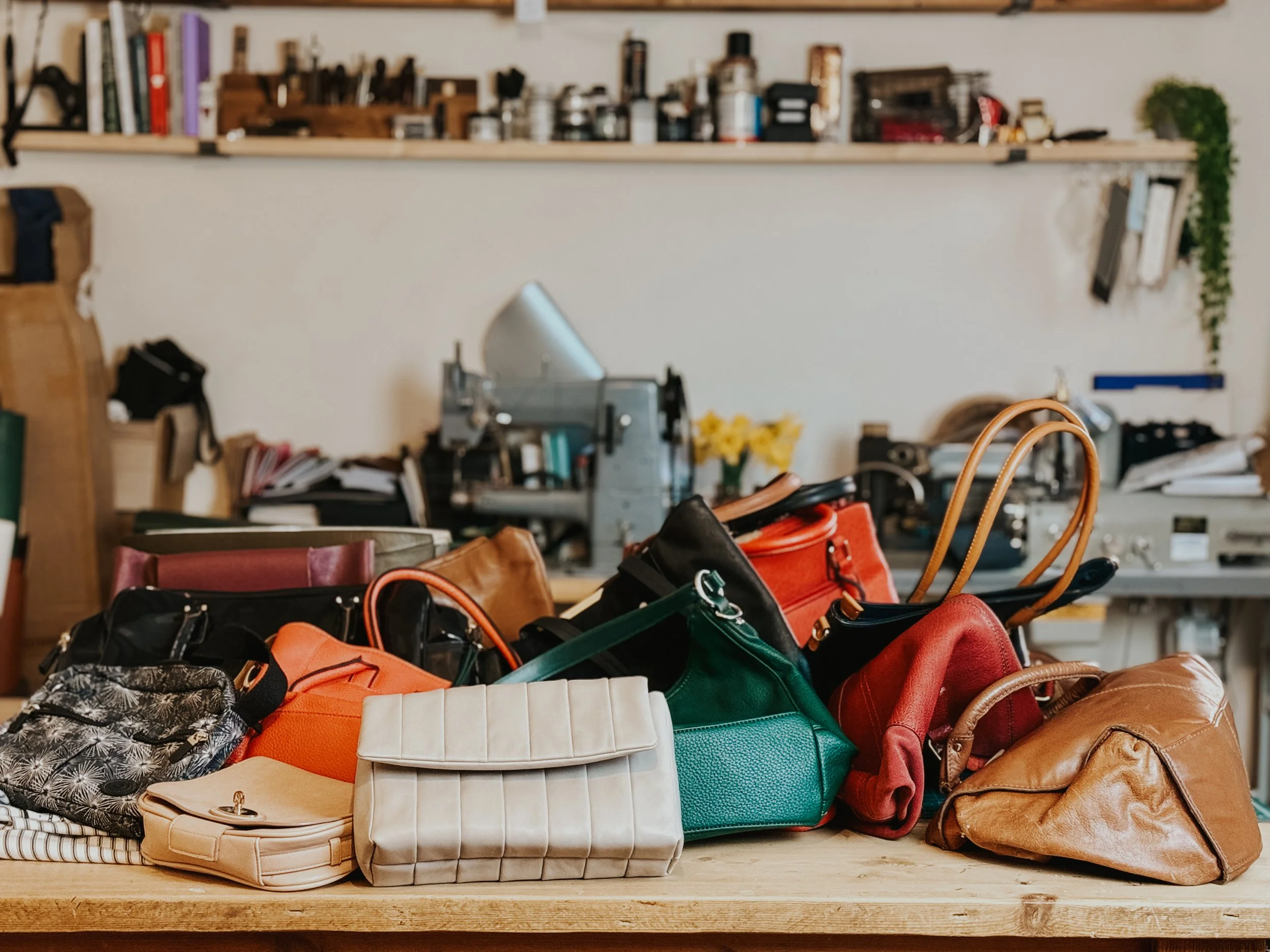 A variety of handbags and purses in different colors and styles on a wooden table in a workshop or studio setting, with tools, books, and plants in the background.