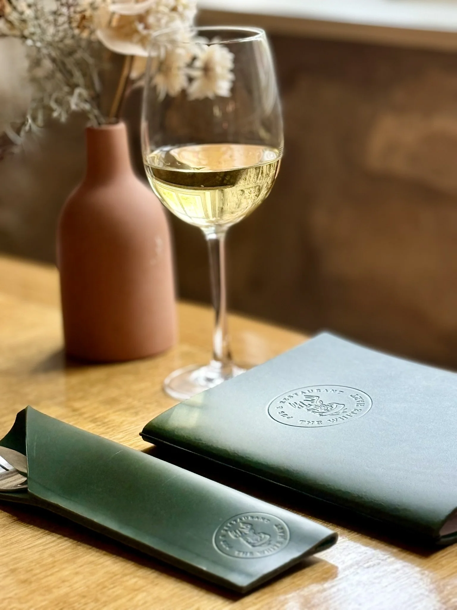 A glass of white wine, two green napkins with a logo, and a pink vase with dried flowers on a wooden table.
