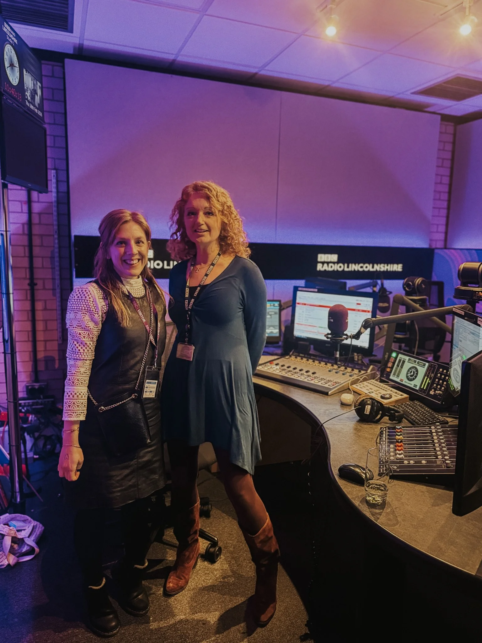 Two women in a radio studio posing for a photo. The studio has a large desk with radio equipment, computers, listening devices, and a microphone. There is a large screen in the background displaying 'BBC Radio Lincolnshire'.