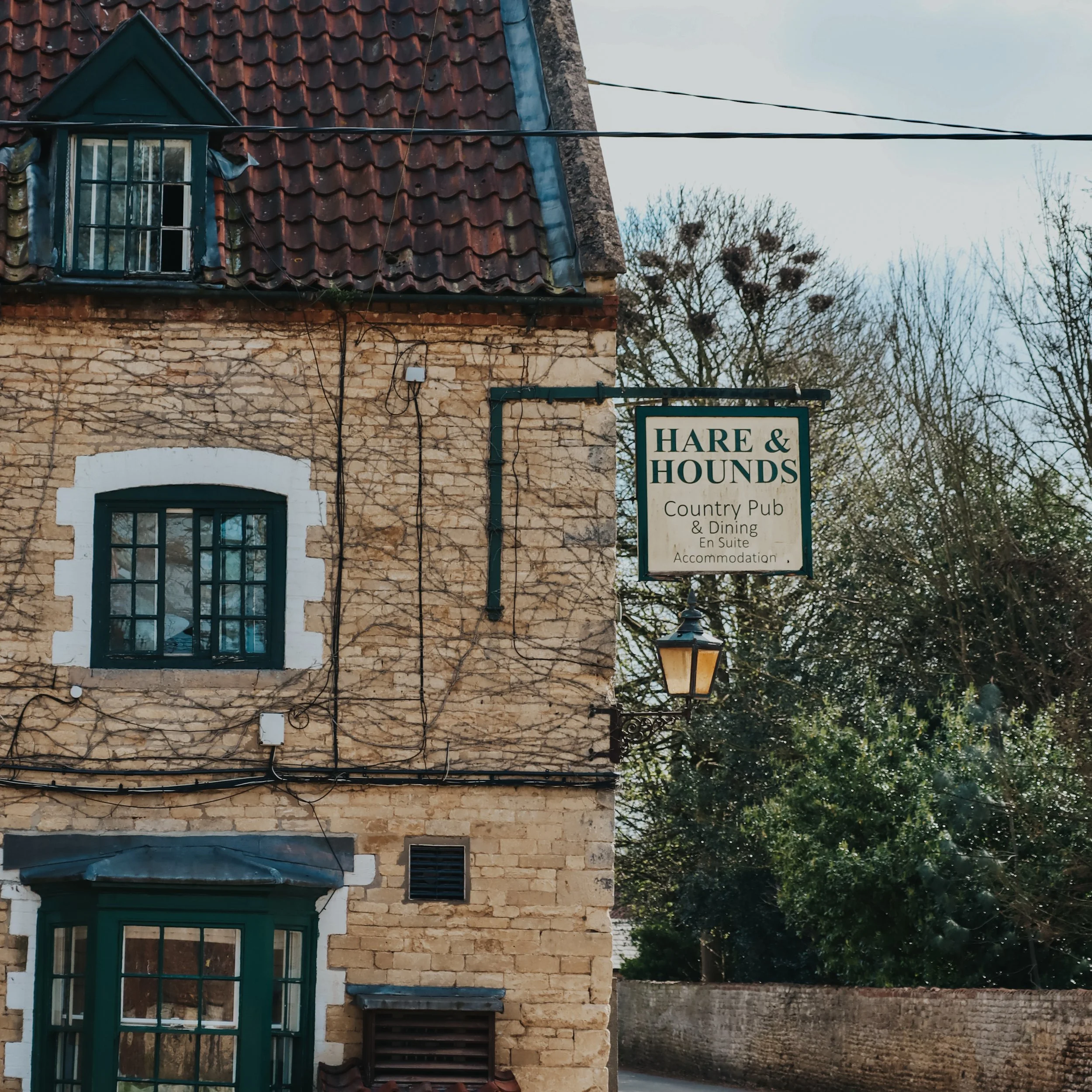 A beige brick building with black-framed windows and a sign that reads 'Hare & Hounds, Country Pub & Dining, En Suite Accommodation'. There is a streetlamp attached to the building and trees in the background.
