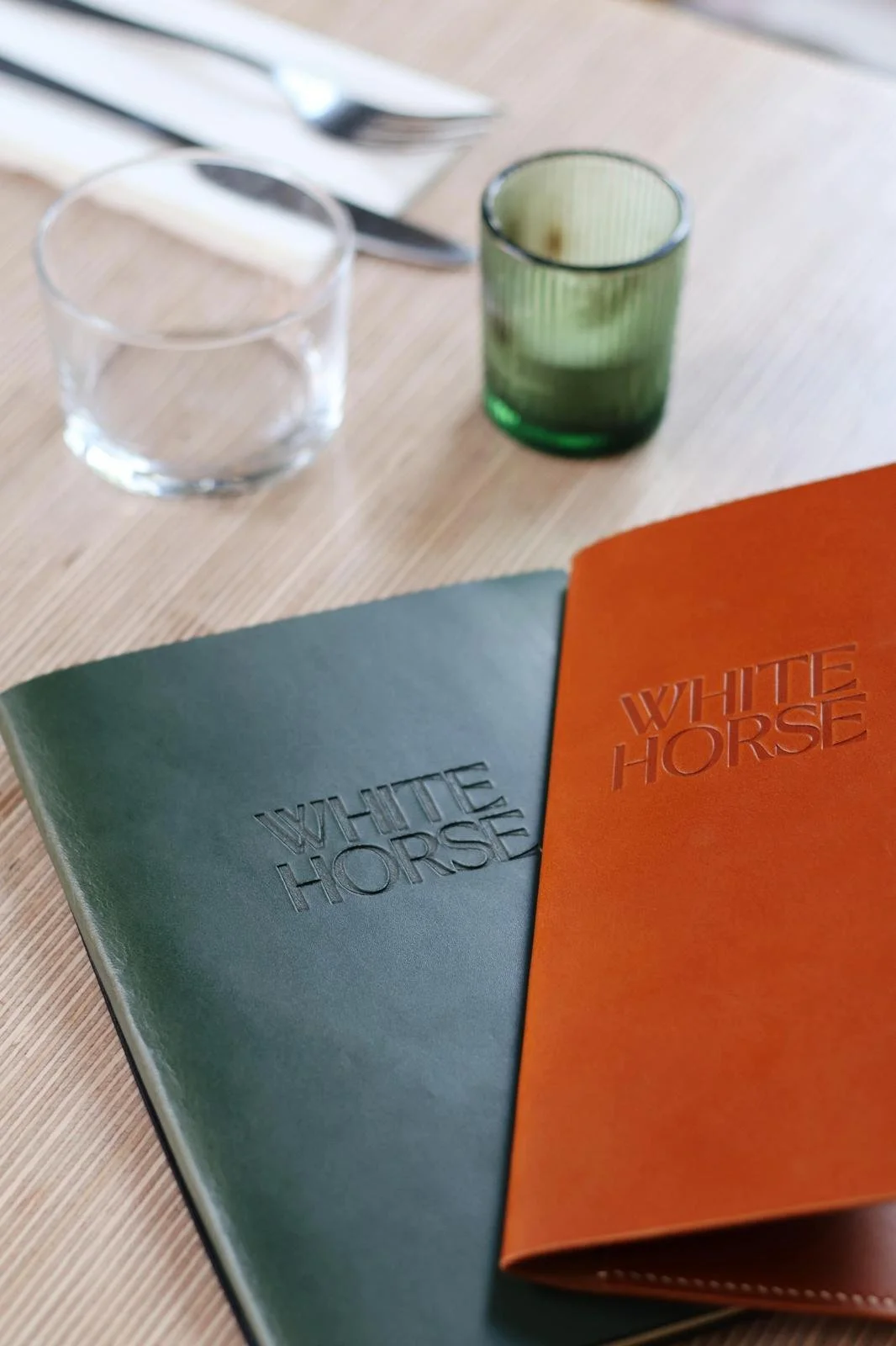 Two menus, one green and one orange, on a wooden table with two glasses, one clear and one green, and cutlery in the background.