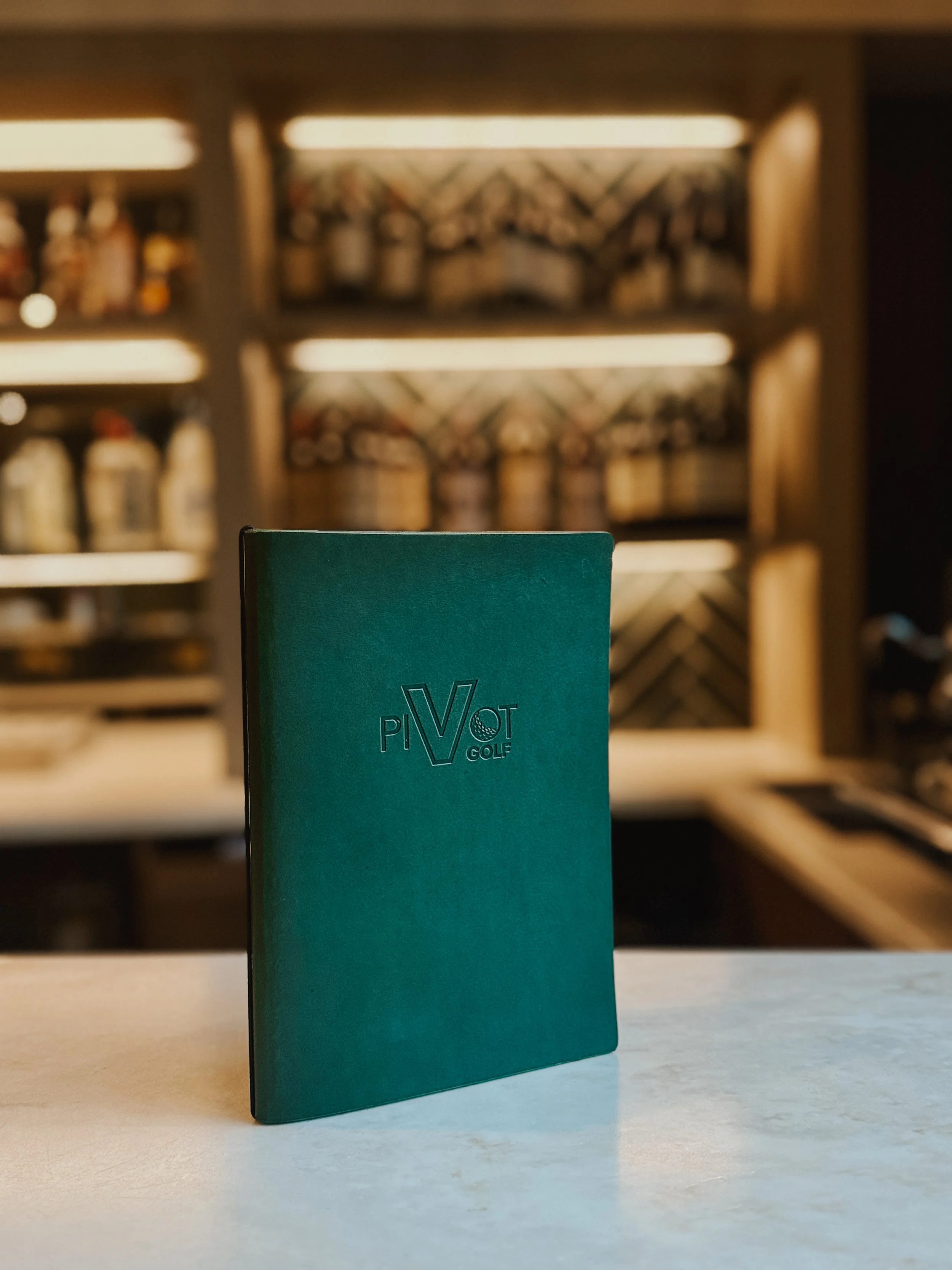 Green menu or booklet with 'PiVOT GOLF' embossed on the cover, standing upright on a white surface, with a blurred background of shelves with bottles and decorative items.