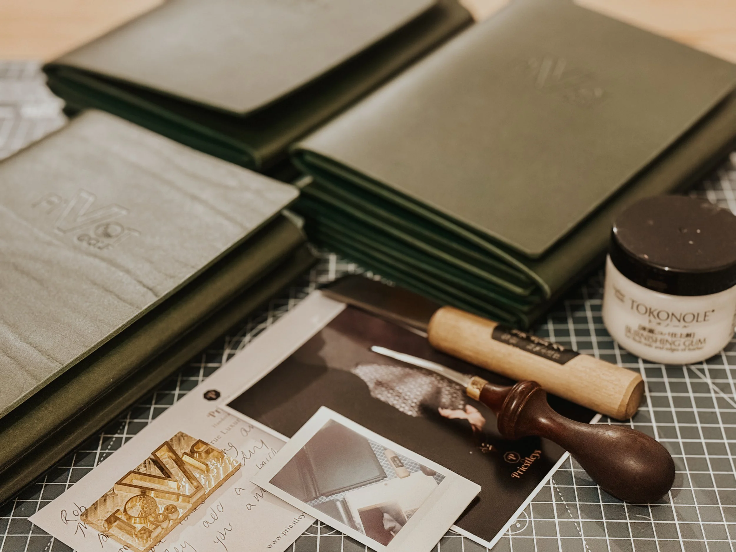 Crafting tools and materials on a cutting mat, including leather wallets, a stitching hammer, a leather carving tool, a jar of leather glue, and photographs of leatherwork.