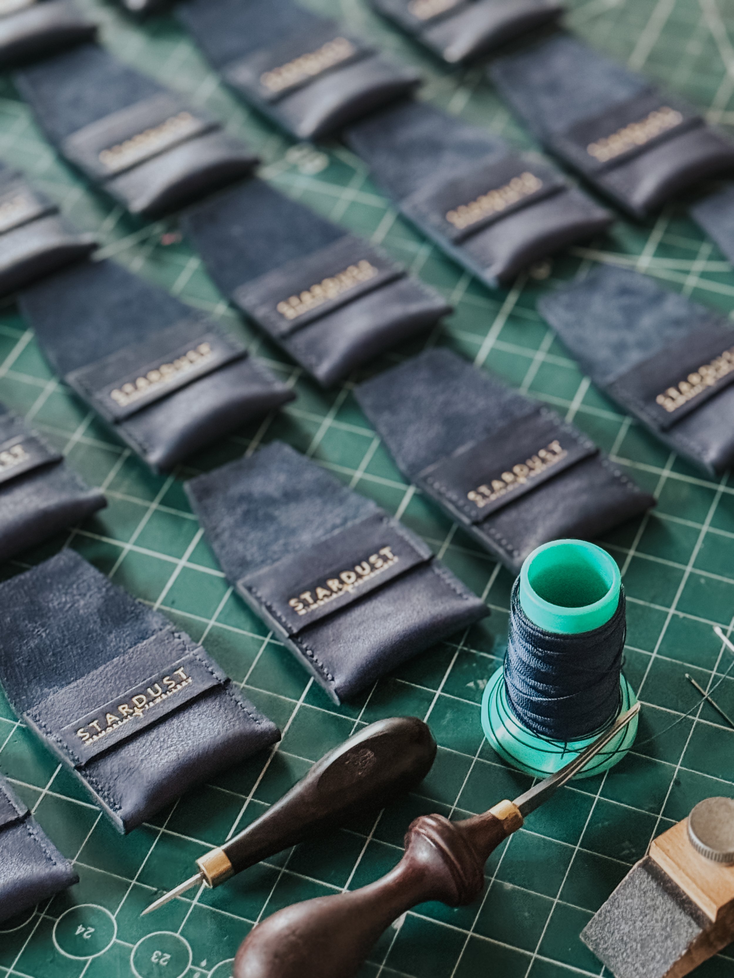 Close-up of multiple black leather watch straps with the brand name 'Starboard' embossed in gold, arranged on a green cutting mat with grid lines. A spool of black thread and a sewing needle are also visible in the foreground.