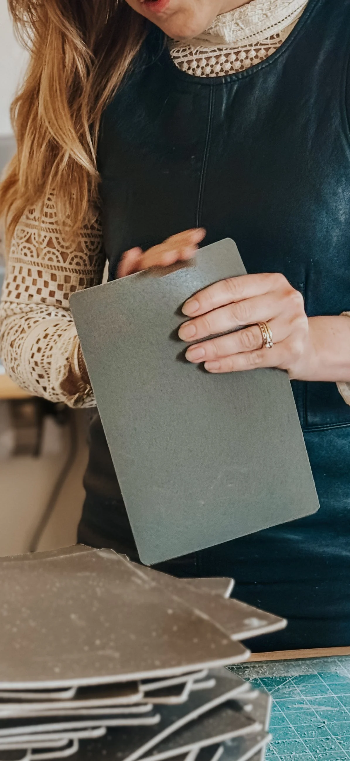 Close-up of a woman wearing a lace blouse and leather vest, holding a gray sheet of paper or cardboard, with jewelry on her ring finger.