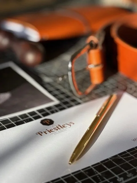 Close-up of a desk with a white notepad labeled 'Prestley,' a gold pen, an orange watch, and glasses on a black grid surface.