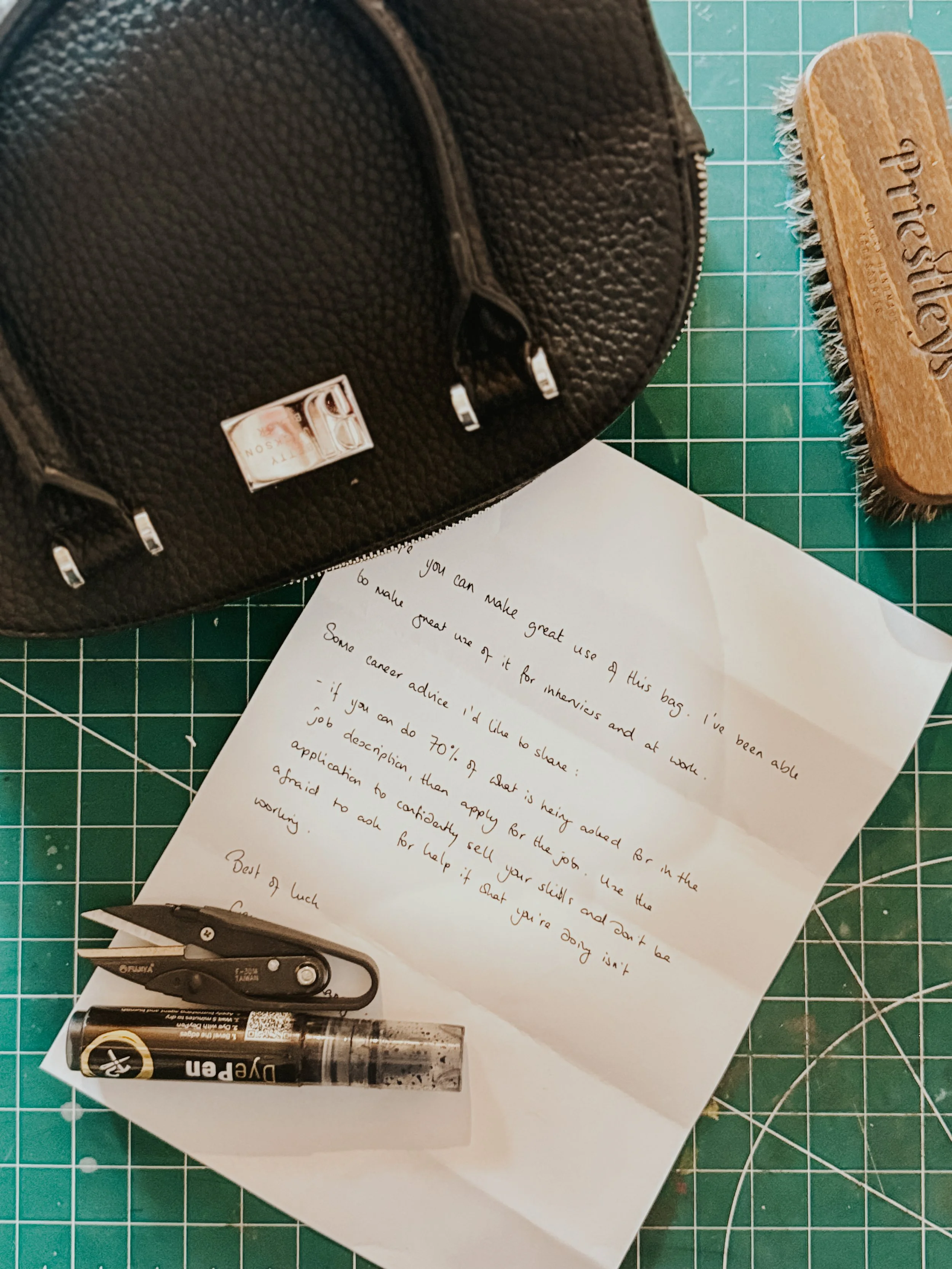 Letter on green cutting mat, black leather bag, wooden shoe brush, black pen, and silver utility knife.