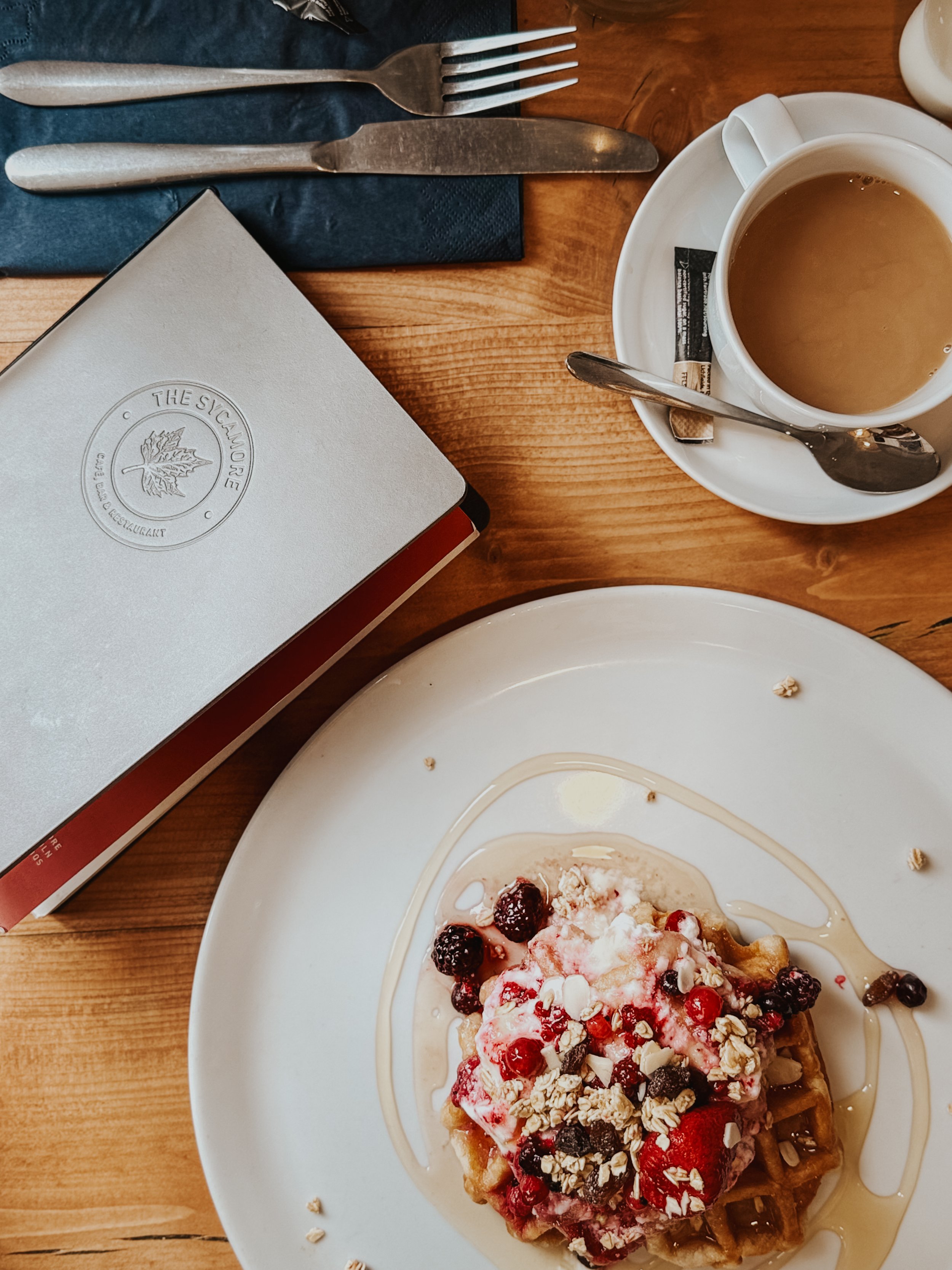 Waffle topped with mixed berries, whipped cream, and sliced almonds on a white plate at a wooden table, with a cup of coffee nearby.