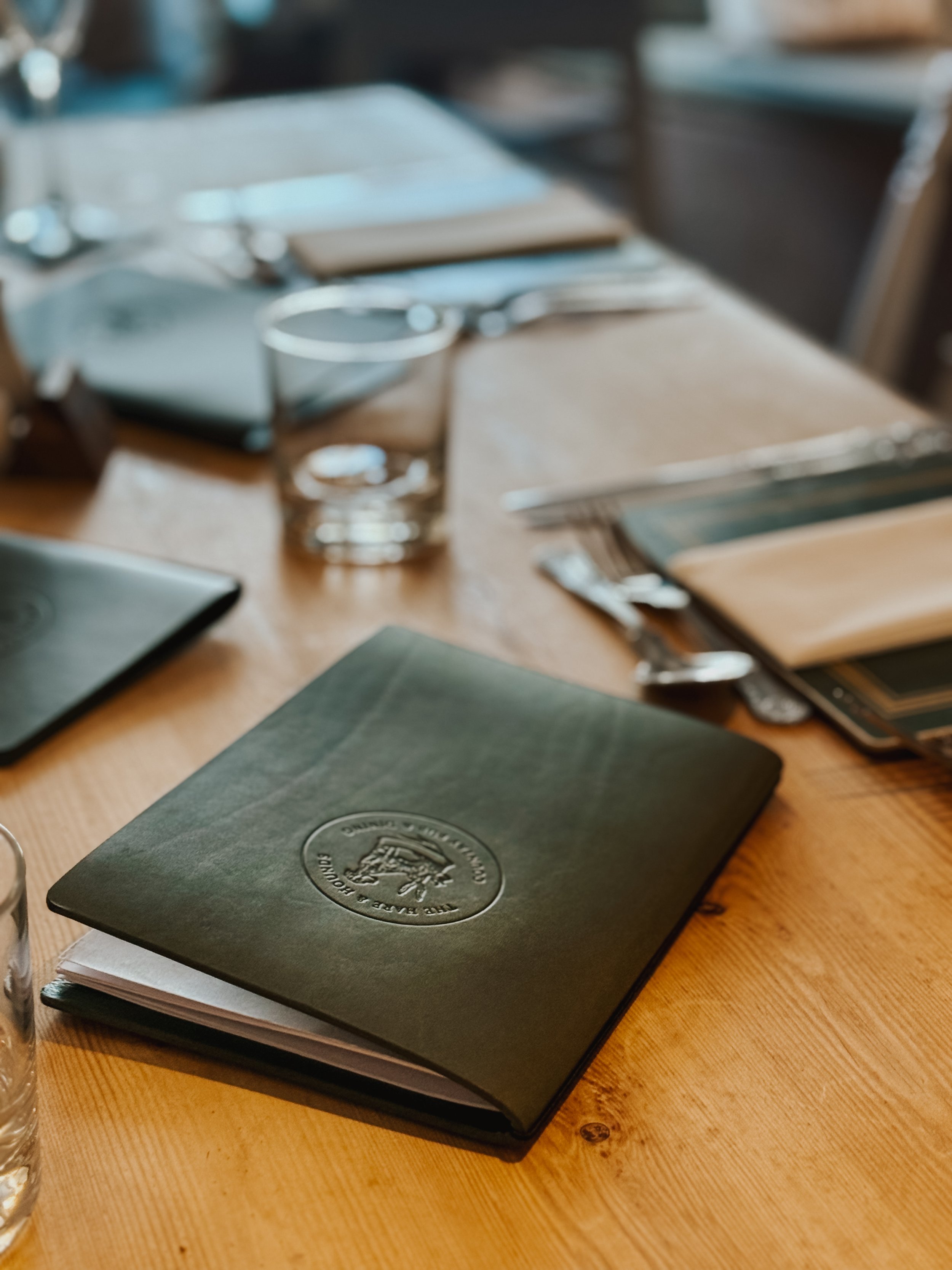 A table with a closed green leather menu, glass of water, and silverware in a restaurant setting.