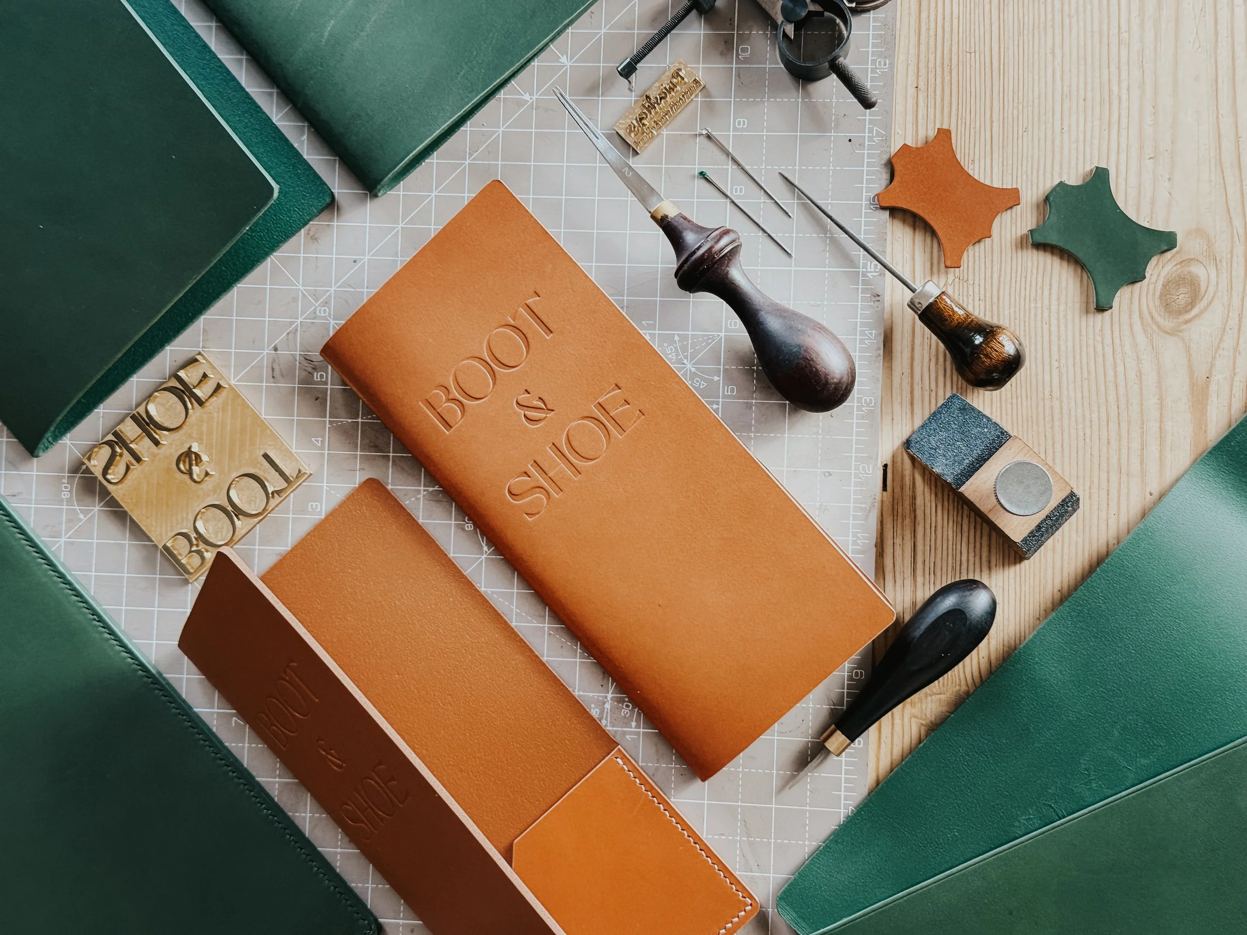 Leather craft tools and materials on a wooden work surface, including leather pieces, a stamping hammer, a swivel knife, and punching tools, with green leather folders and a ruler grid.