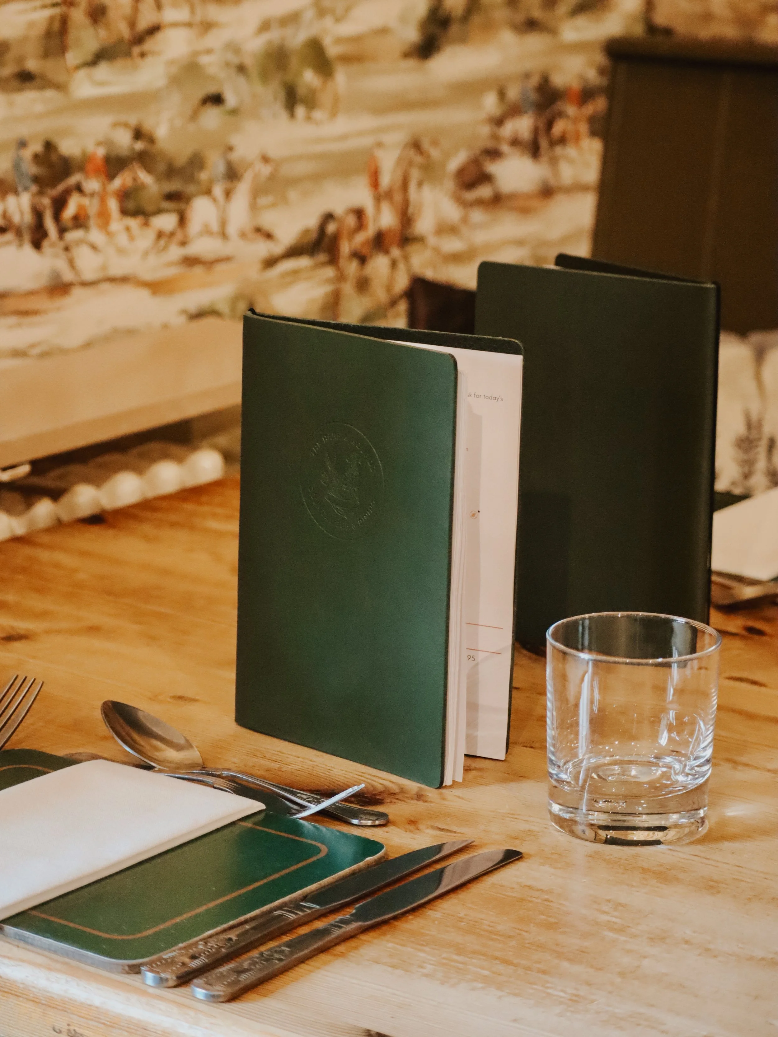 A wooden table in a restaurant setting with a green menu, black napkin, clear glass, utensils, and a white cloth napkin.