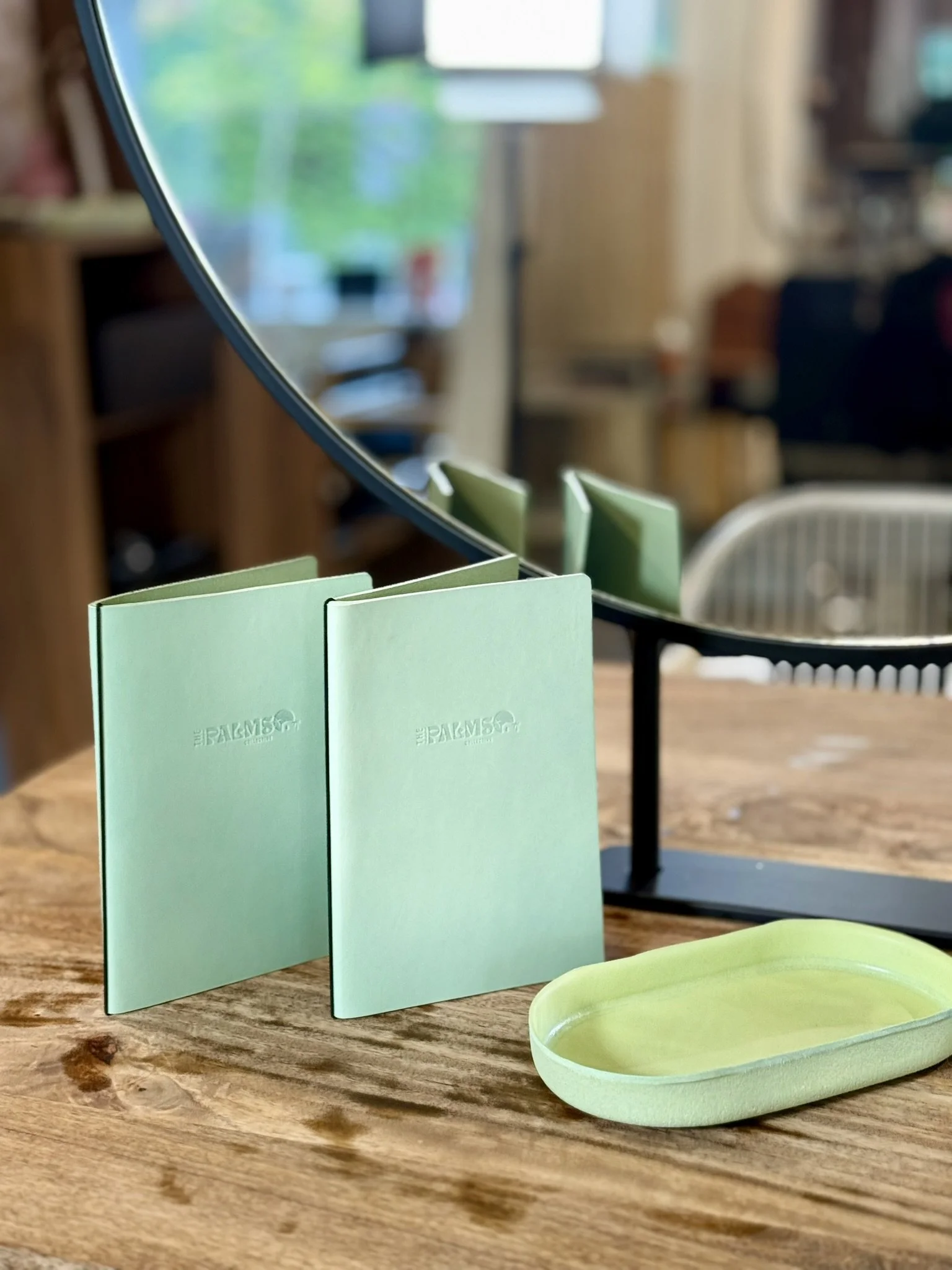 A mirror with green sticky notes attached to its frame, a small green dish, and a wooden table in a cozy room with blurred background furniture and window.