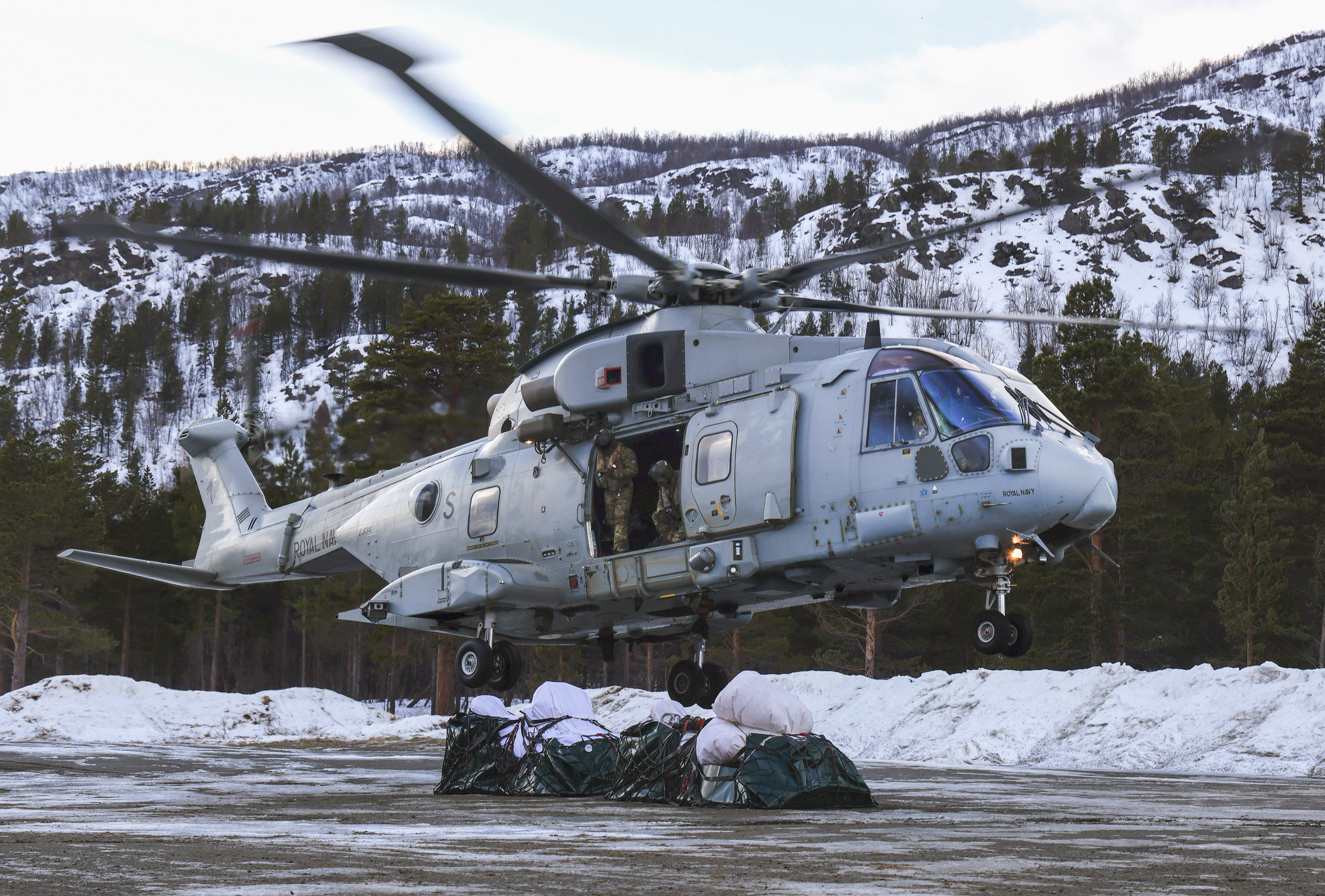 A military helicopter is hovering above a snow-covered ground in a mountainous area with trees. Soldiers are standing near the opening in the helicopter.