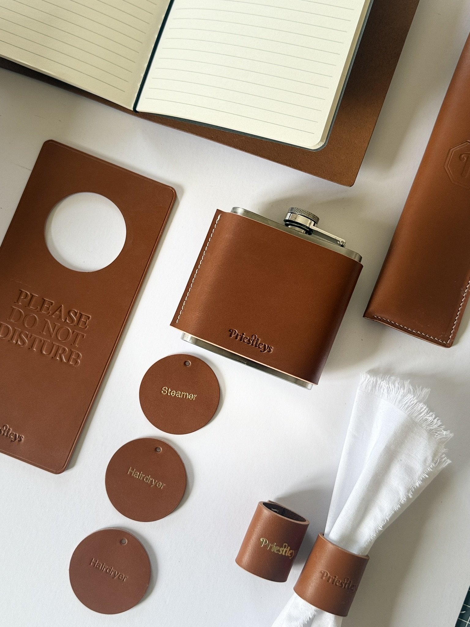Flat lay of various brown leather items including notebooks, a flask with a cap, round tags labeled 'Steamer' and 'Hairdryer,' and a white cloth, all arranged on a white surface.