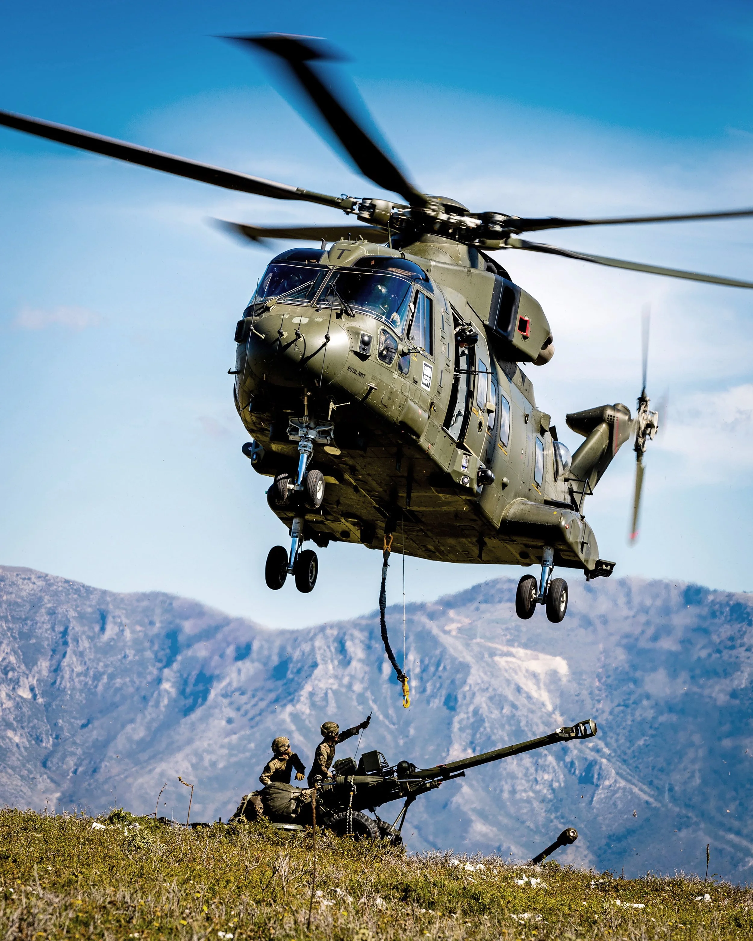 A military helicopter flying low over soldiers operating an artillery gun on a grassy field with mountains in the background.