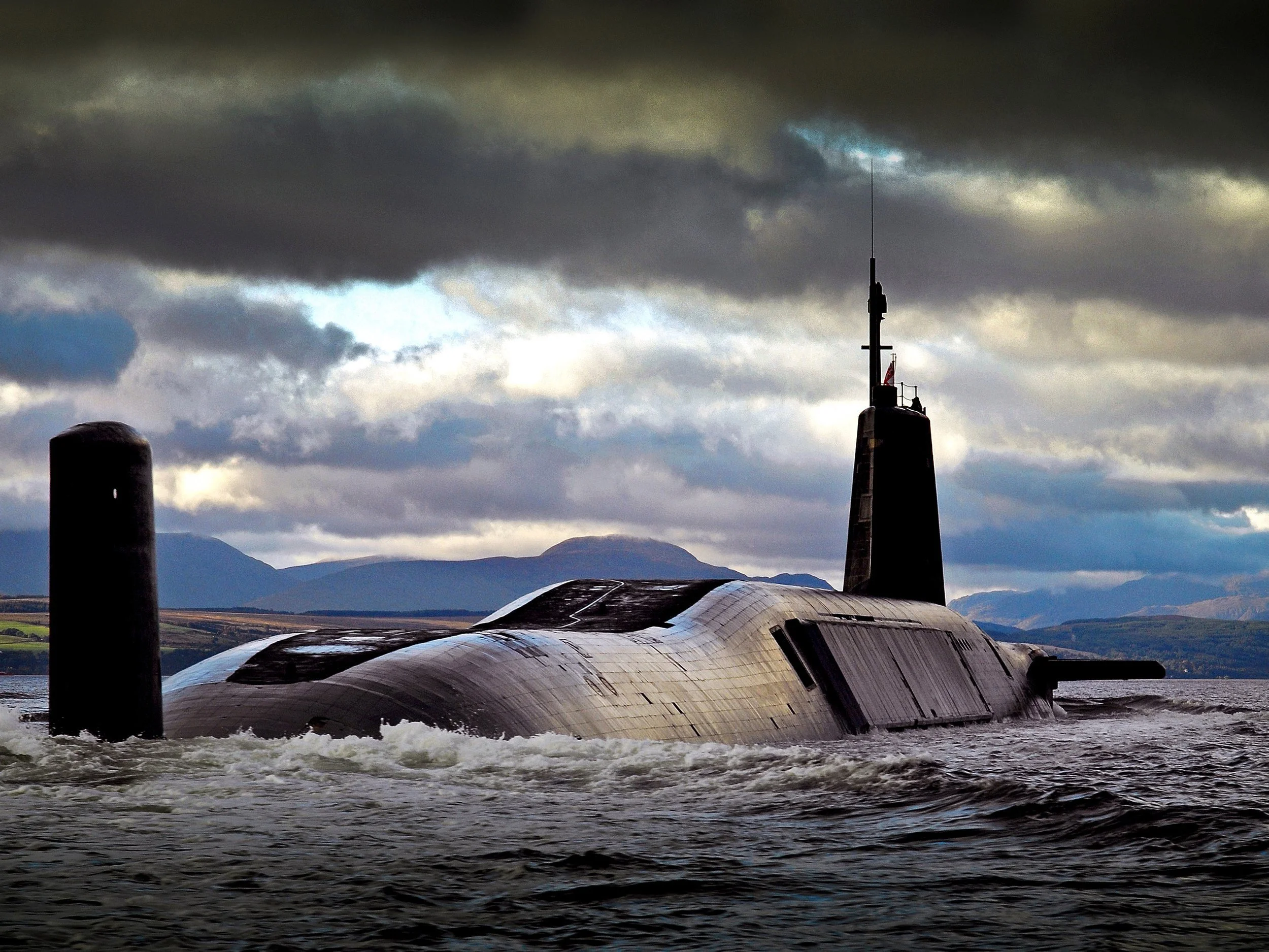 A submarine partially submerged in water with mountains and a cloudy sky in the background.