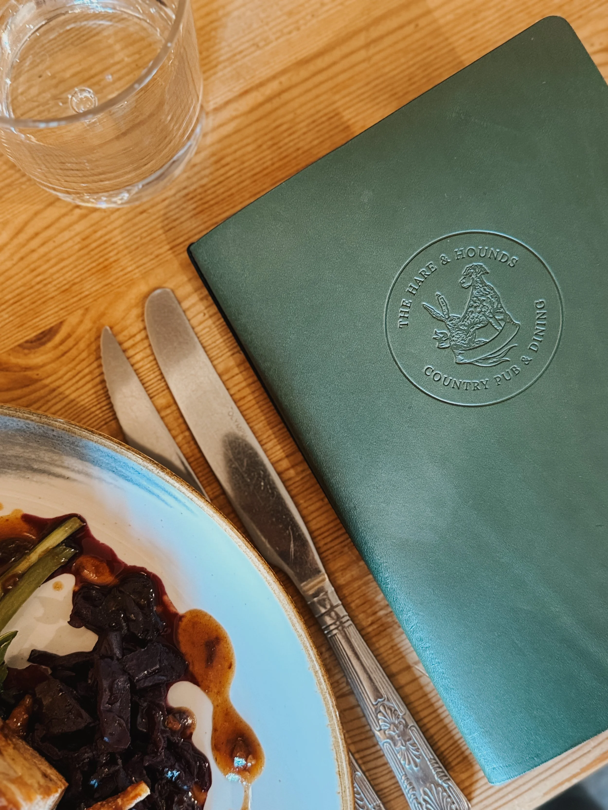 A place setting at a restaurant including a glass of water, a green menu with the logo 'The Hare & Hounds Country Pub & Dining,' a knife and fork, and part of a plate with food on it.