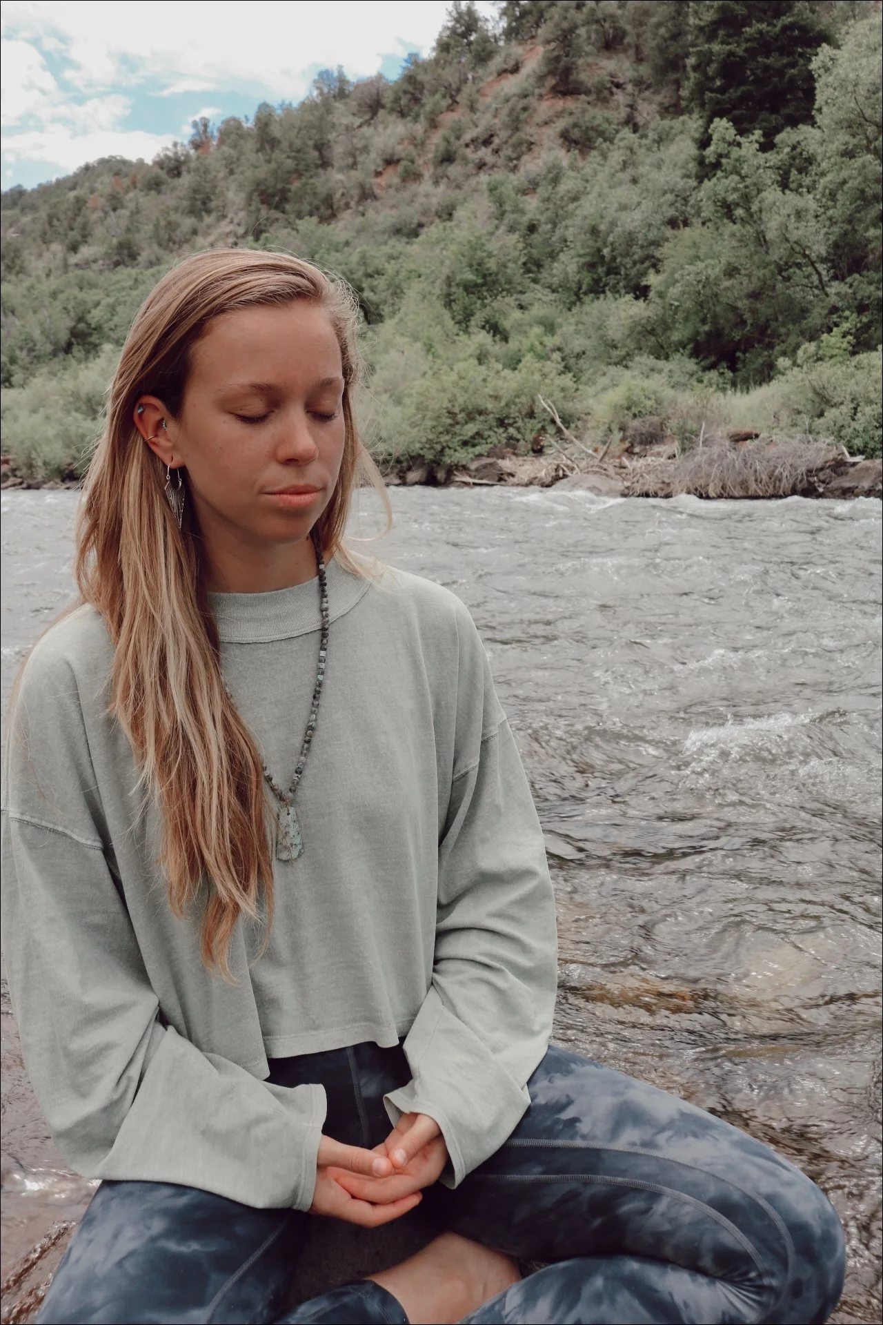 A woman sitting by a river with her eyes closed, wearing a gray crop top, black pants, a beaded necklace, and earrings, with a forested hillside in the background.