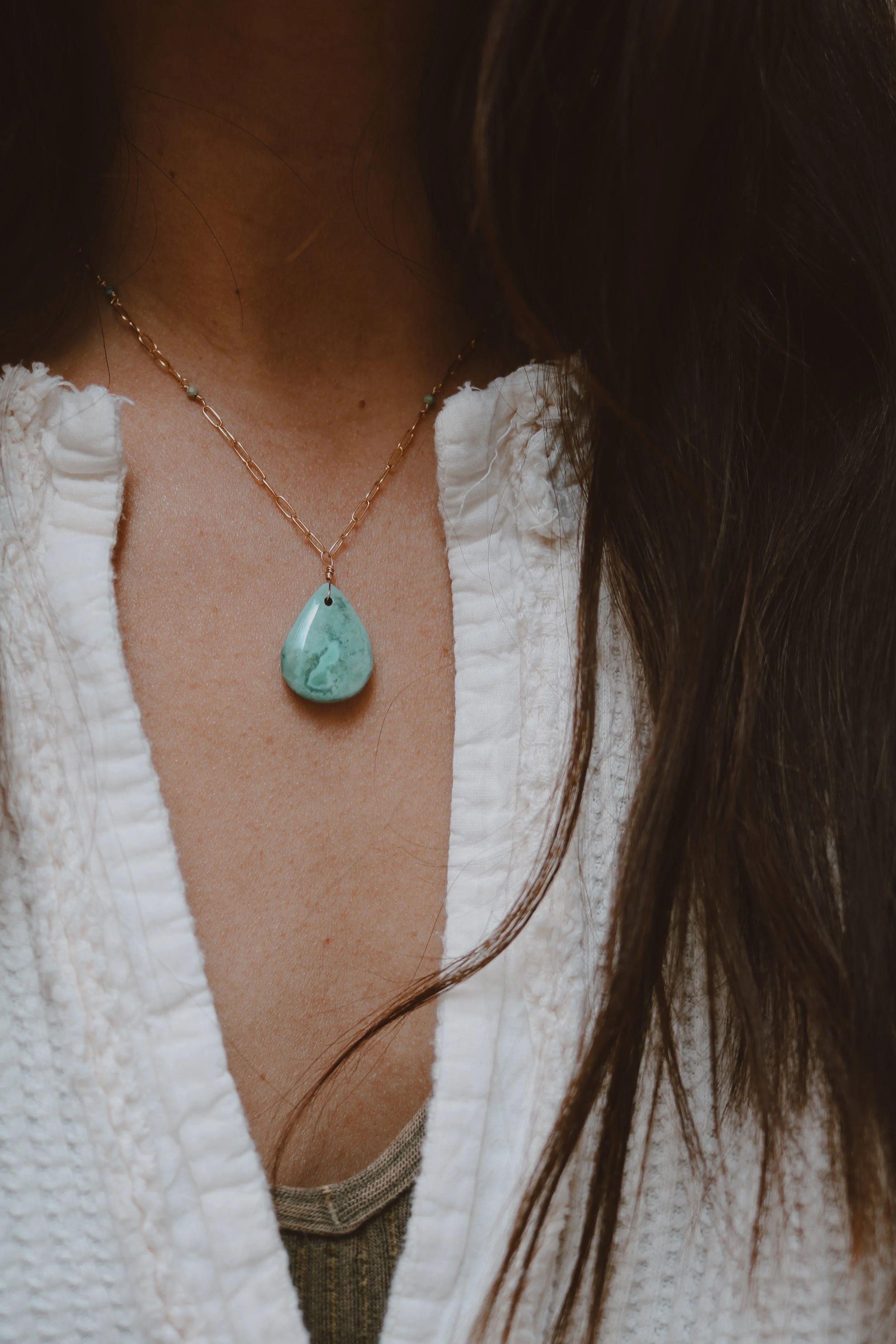 Close-up of a woman wearing a necklace with a large greenish-blue pendant, partially visible white textured shirt, and long brown hair.